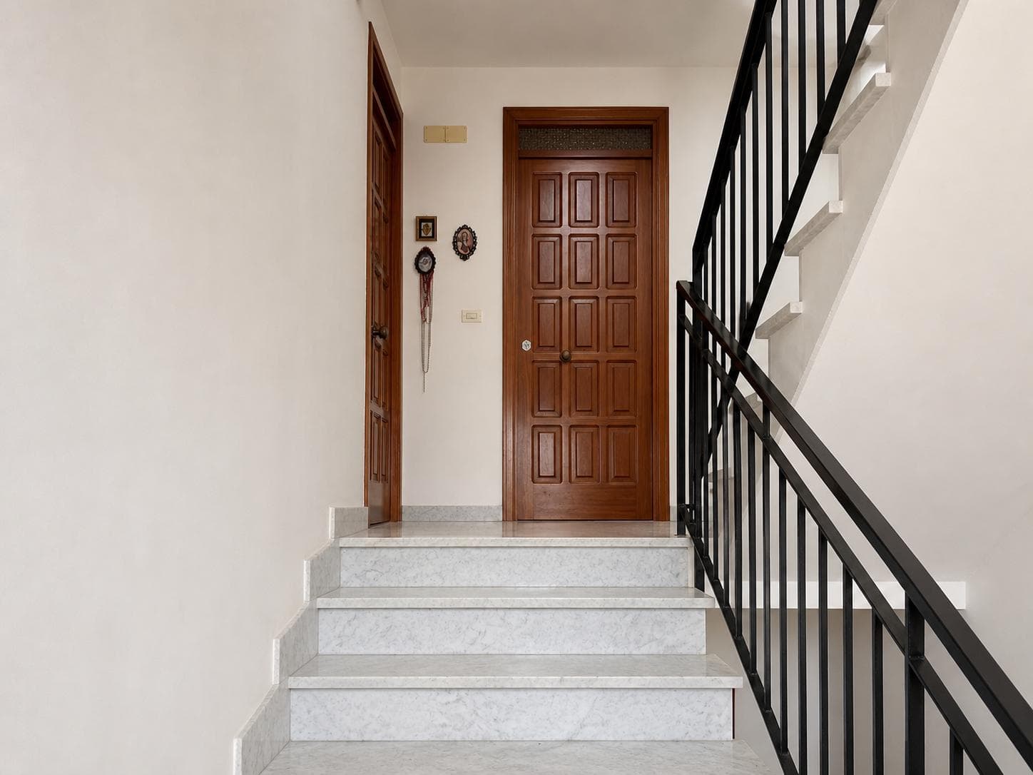 Porta d'ingresso in legno massiccio con dettagli classici, situata in un ingresso luminoso e accogliente, con pareti bianche e decorazioni tradizionali. Perfetta per case che cercano stile e funzional.