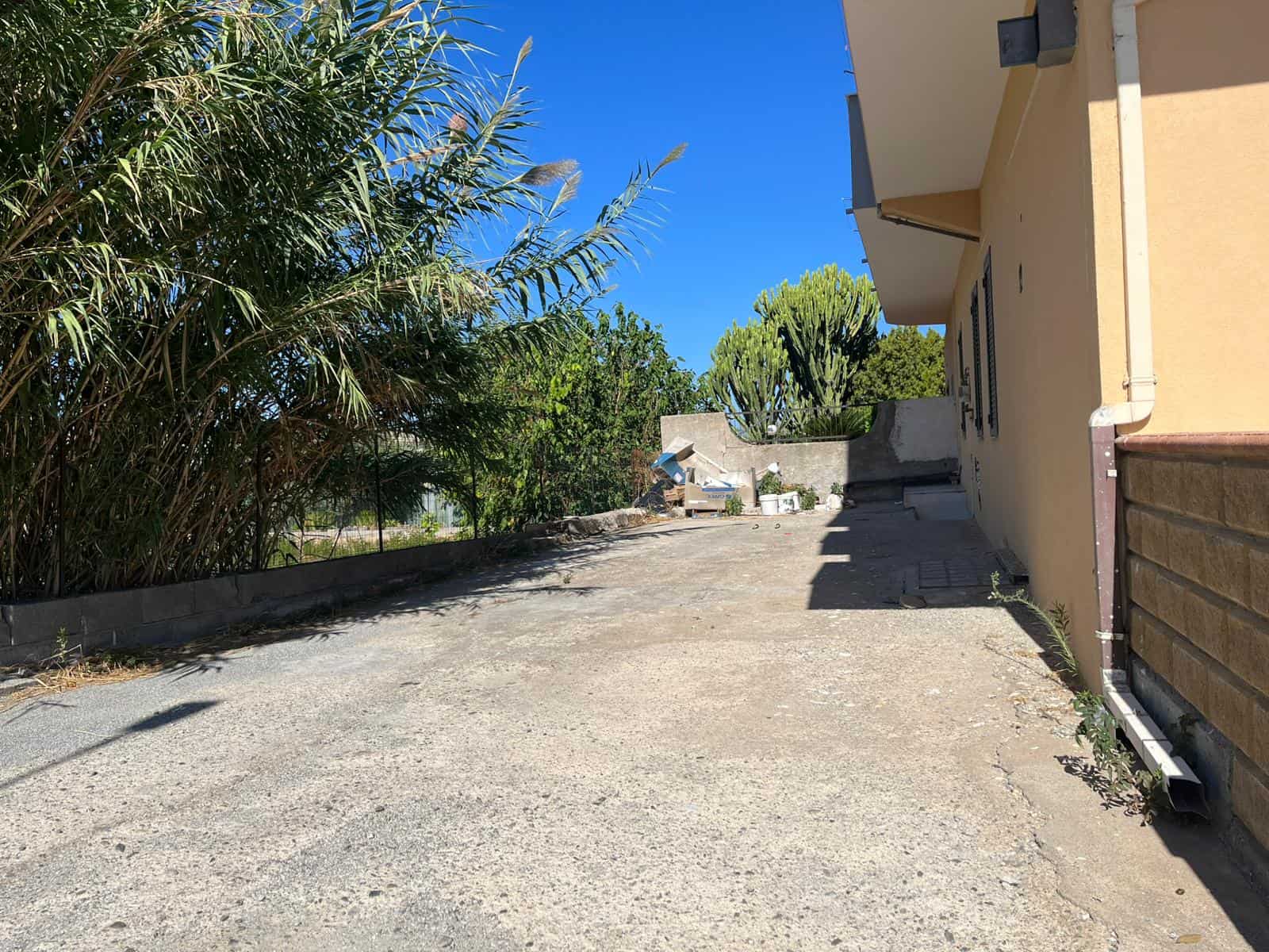 Ampia area esterna con spazio di parcheggio e vegetazione lussureggiante, vicino a una casa residenziale in zona tranquilla e soleggiata, ideale per attività all'aperto e relax.