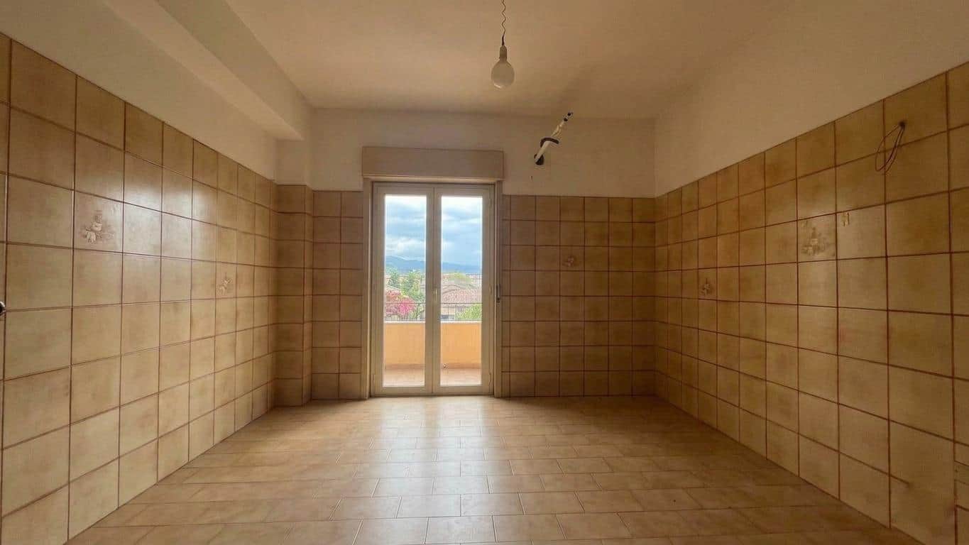 Ampio soggiorno vuoto con pareti rivestite di piastrelle beige e grande finestra con vista panoramica sulla città e le colline circostanti.