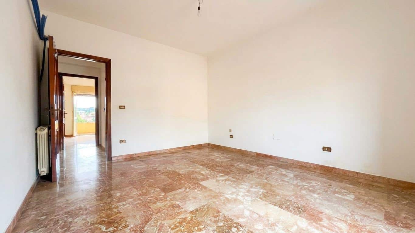 Ampia stanza vuota con pareti bianche e pavimento in marmo, luce naturale che entra da una porta aperta che conduce a un balcone con vista panoramica. Ideale per ristrutturazione e personalizzazione.