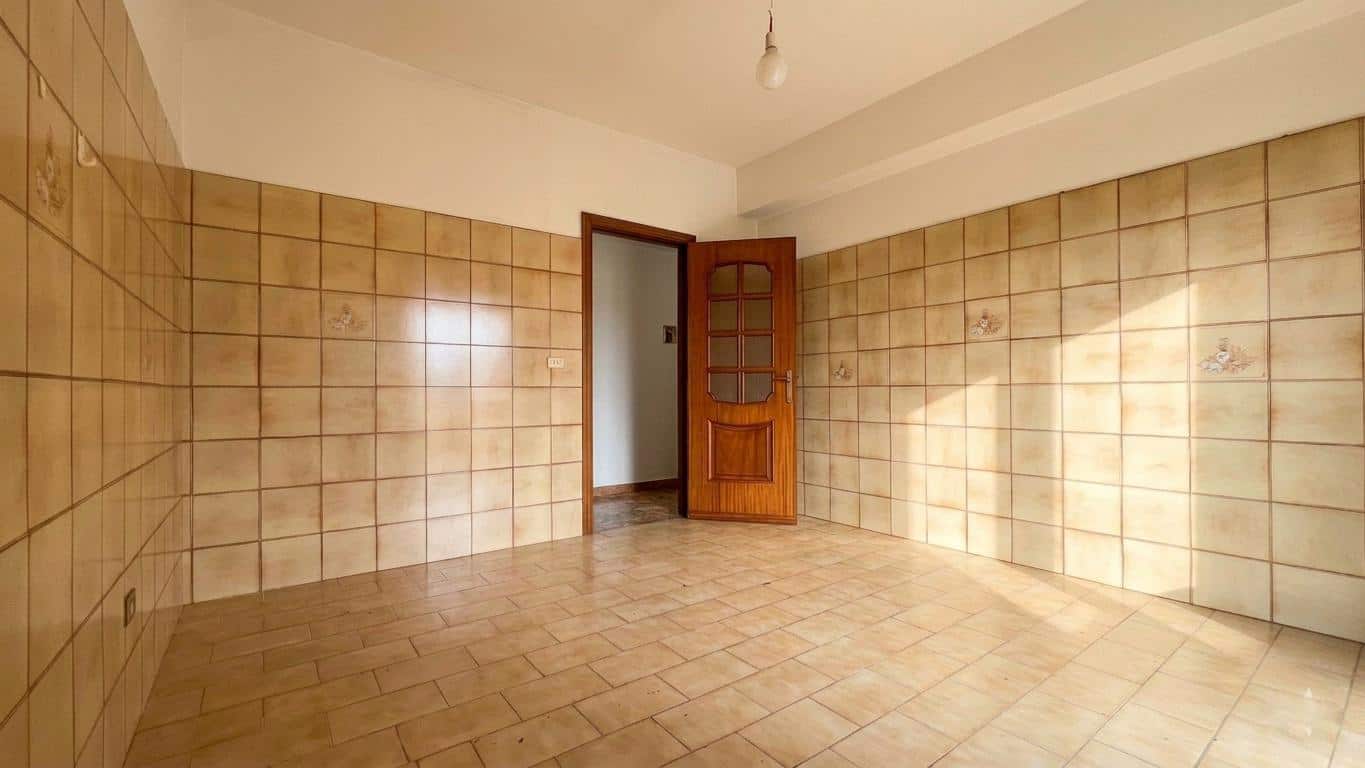 Ampia stanza vuota con pareti rivestite di piastrelle beige e porta in legno aperta, illuminata dalla luce naturale.