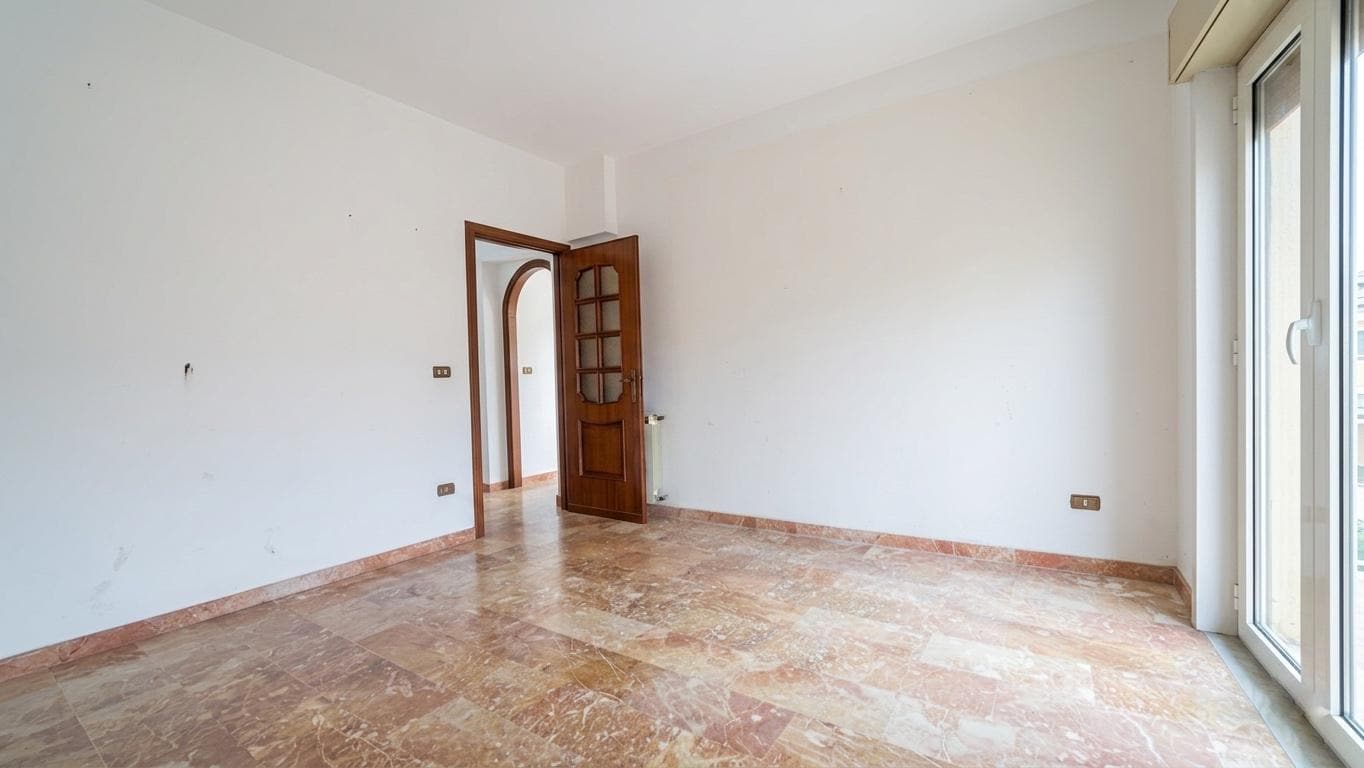 Ampia stanza vuota con pareti bianche e pavimento in marmo, luminosa grazie a una grande porta finestra che dà su un balcone. Ideale per arredare e personalizzare la propria casa.