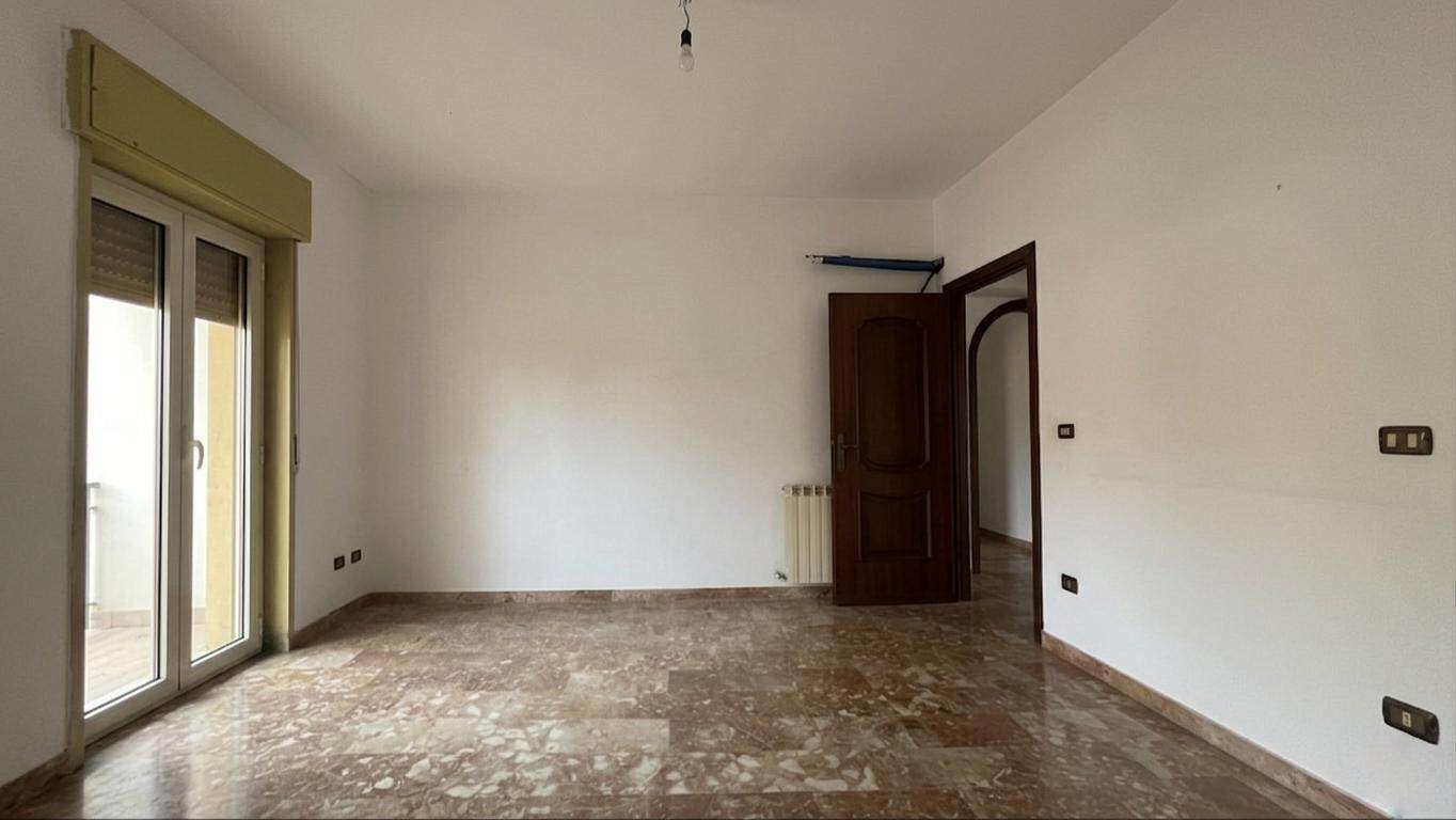 Ampio soggiorno vuoto con pareti bianche, pavimento in marmo e grande porta finestra che dà su un balcone, ideale per ristrutturazione e arredamento personalizzato.