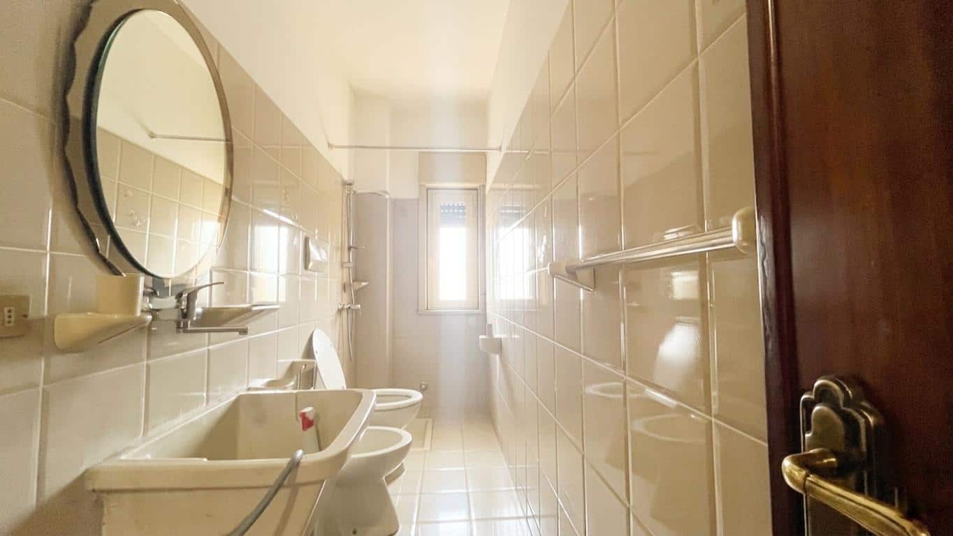 Spazio bagno luminoso con piastrelle beige, lavabo, specchio rotondo e finestra che permette l'ingresso di luce naturale, ideale per ristrutturazioni e miglioramenti domestici.