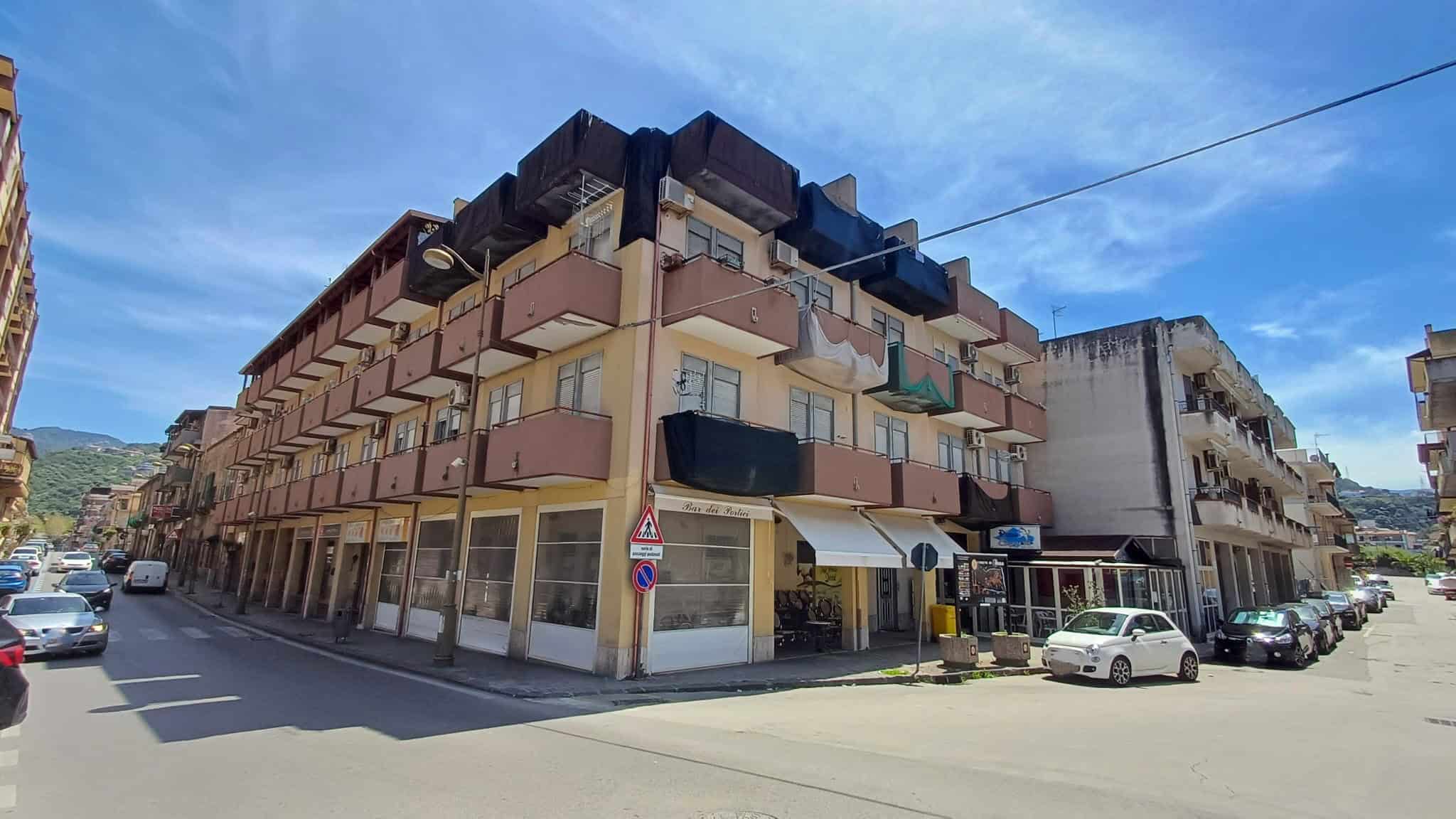 Uscita di un edificio residenziale con negozi al piano terra, situato in un'area urbana soleggiata con veicoli parcheggiati e strade trafficate, in un contesto di città mediterranea.