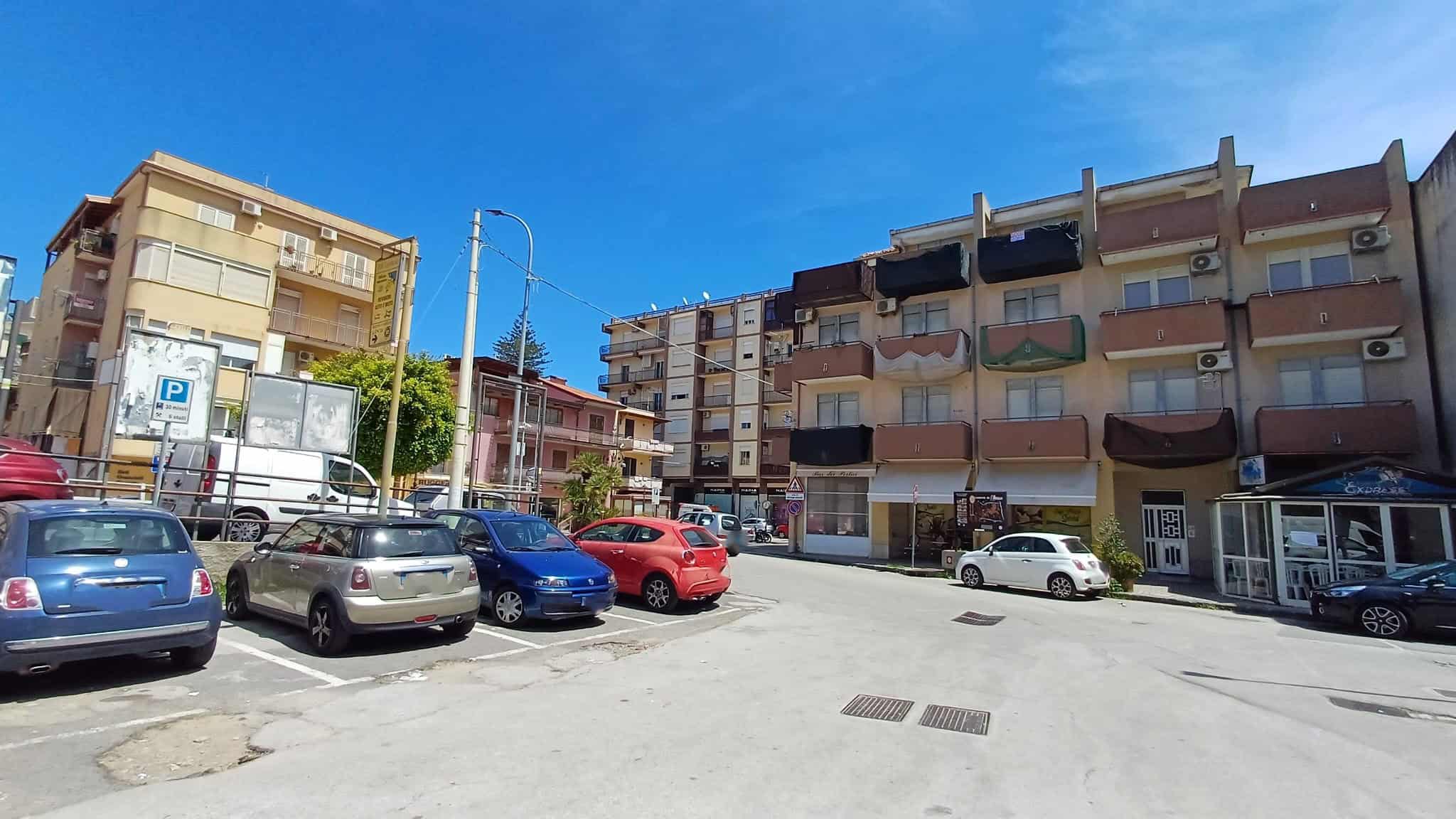 Parcheggio all'aperto davanti a un edificio residenziale in una giornata soleggiata, con auto parcheggiate e un cielo azzurro.