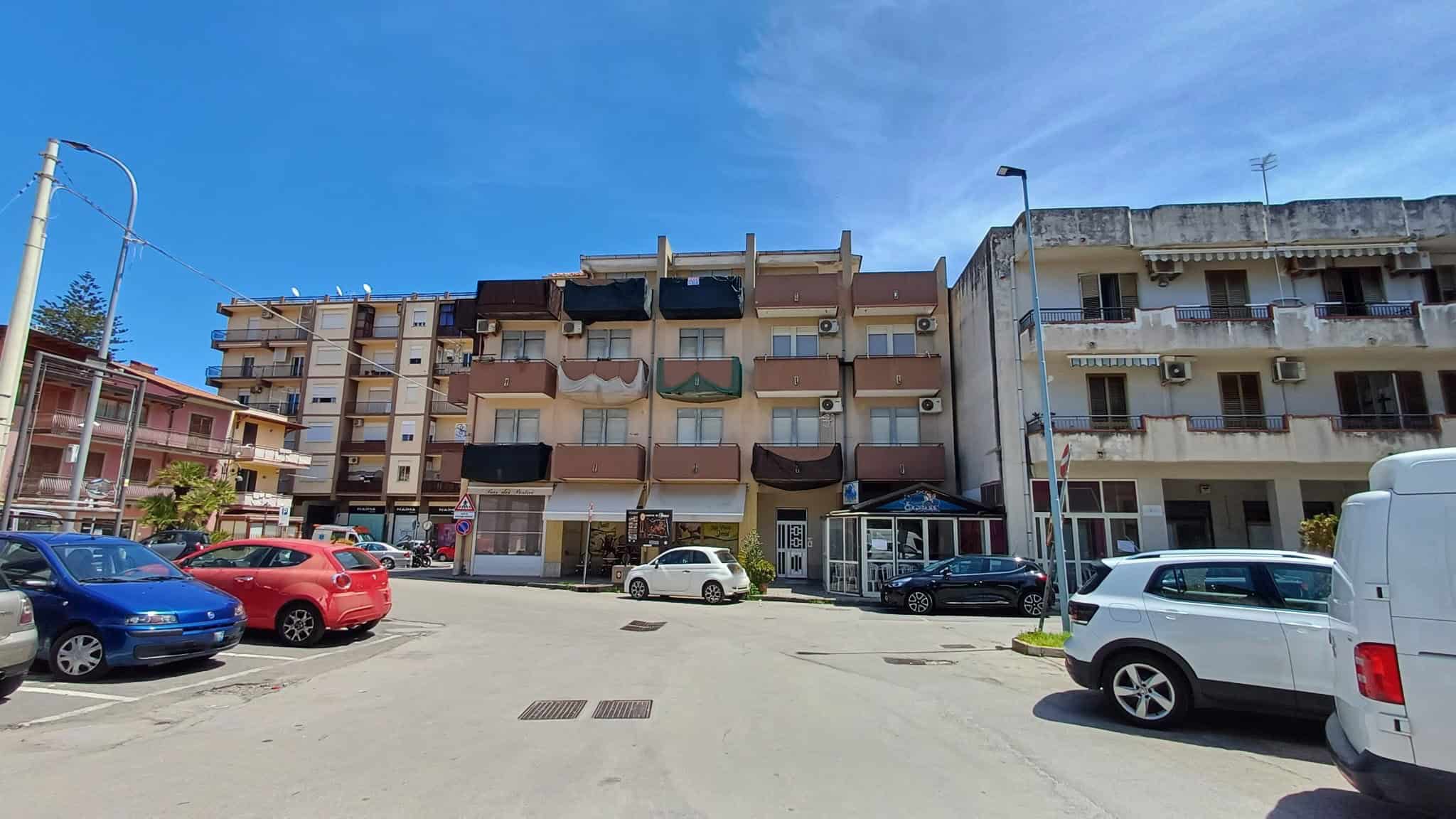 Appartamenti residenziali in un edificio multifamiliare con balconi, situato in una zona urbana con parcheggi e strada trafficata, sotto un cielo sereno.