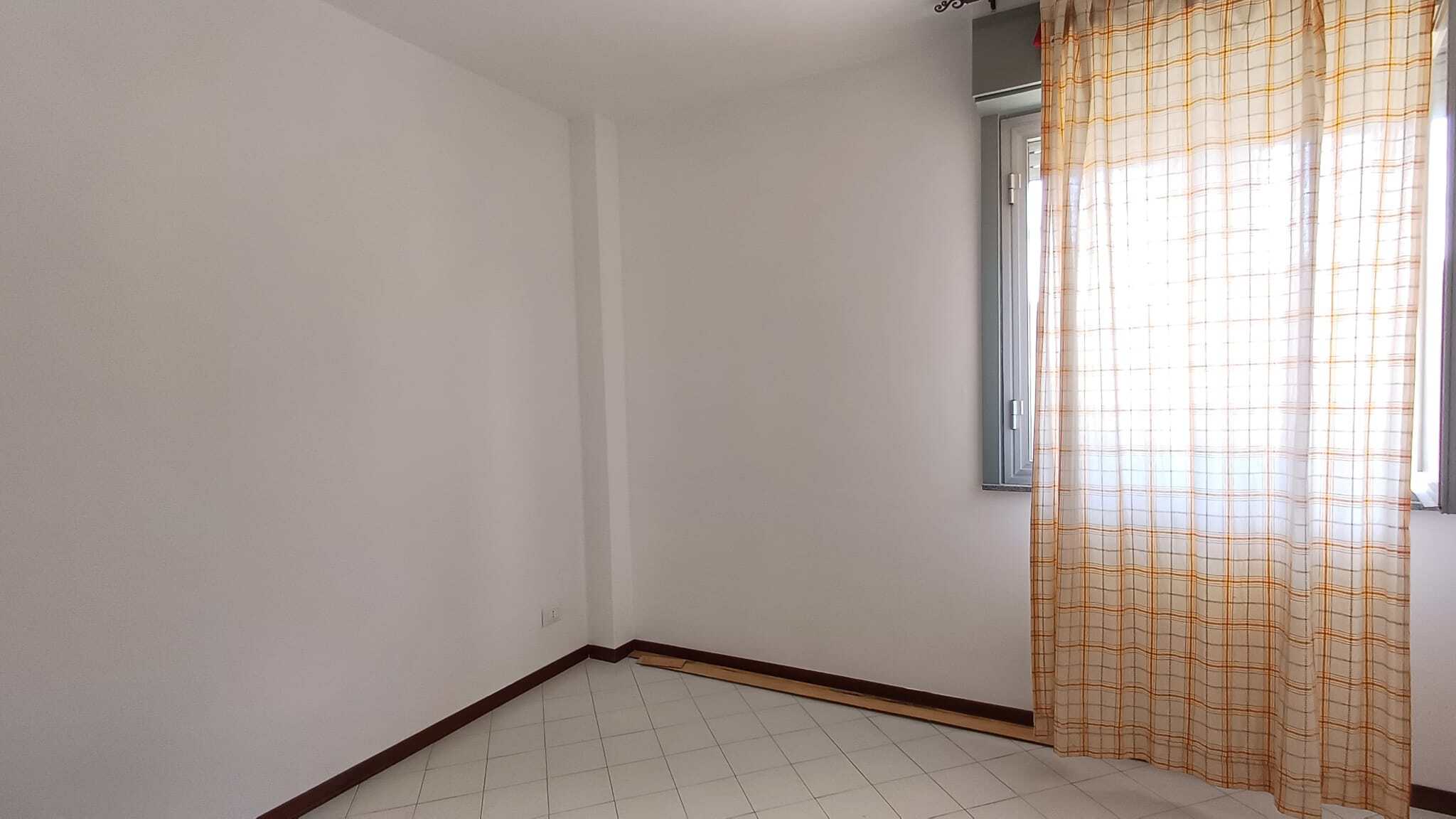 Pannello di finestra con tende a quadretti in una stanza vuota e luminosa, pareti bianche e pavimento in piastrelle.
