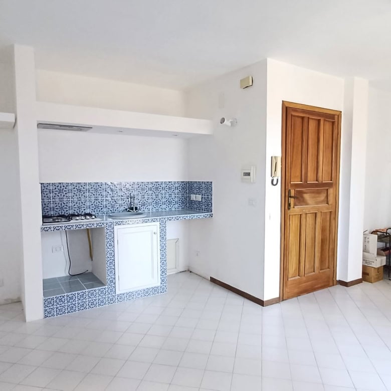 Ampia zona living con cucina a vista, pavimento in piastrelle bianche e pareti neutre, porta in legno e mobili minimalisti, ambiente luminoso e accogliente.