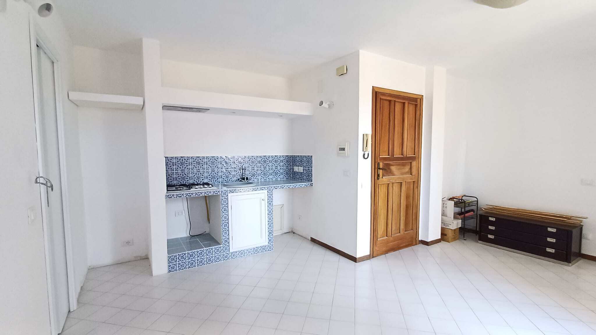 Ampia zona living con cucina a vista, pavimento in piastrelle bianche e pareti neutre, porta in legno e mobili minimalisti, ambiente luminoso e accogliente.