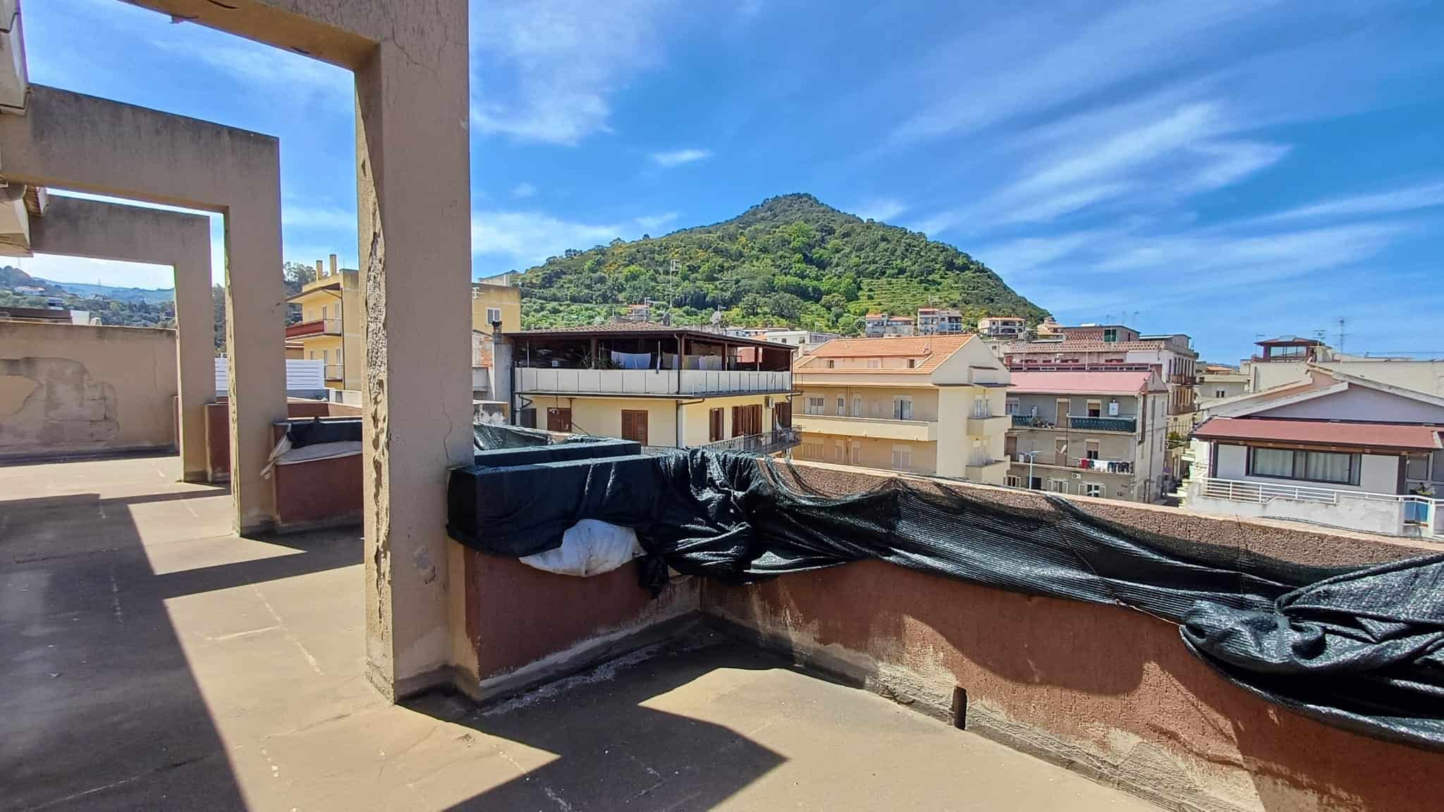 Balcone di un appartamento con vista su un paesaggio urbano e una collina verde, sotto un cielo azzurro con nuvole sparse, in un contesto residenziale italiano.