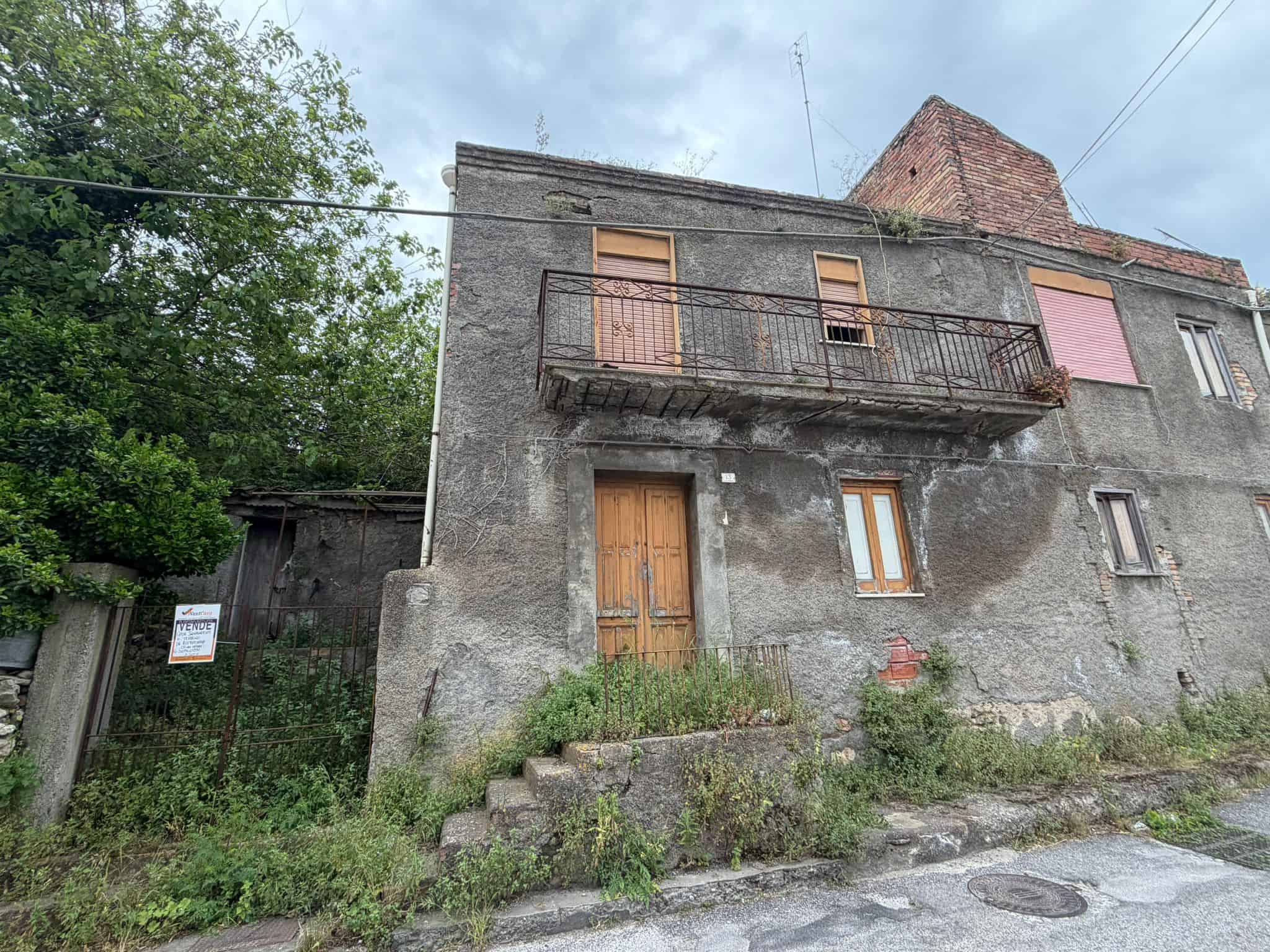 Balcone in ferro battuto e porta in legno su edificio storico in pietra, situato in zona residenziale con vegetazione circostante e strada asfaltata.