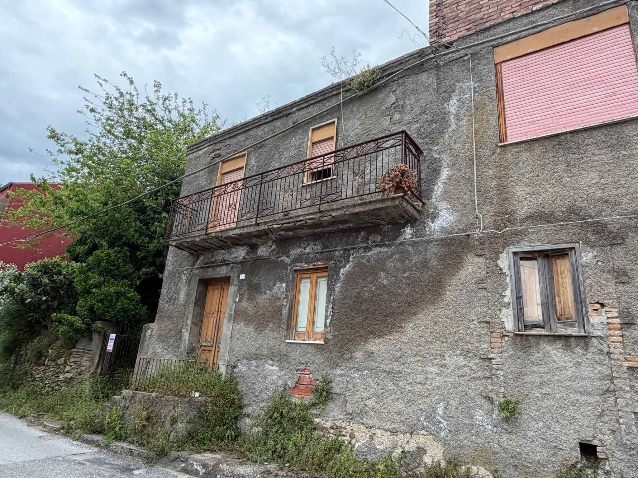 Vecchio edificio residenziale in pietra con balcone in ferro battuto e finestre in legno, situato in un'area rurale con vegetazione e strada sterrata, in condizioni di abbandono.