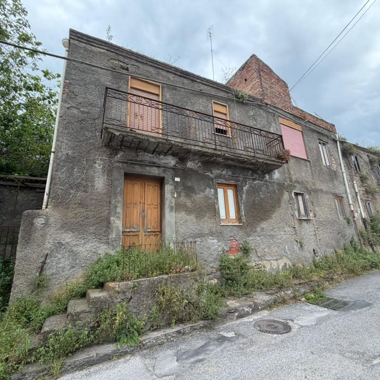 Vecchio edificio residenziale in pietra con balcone in ferro battuto e finestre in legno, situato in zona rurale con strada asfaltata e vegetazione circostante.