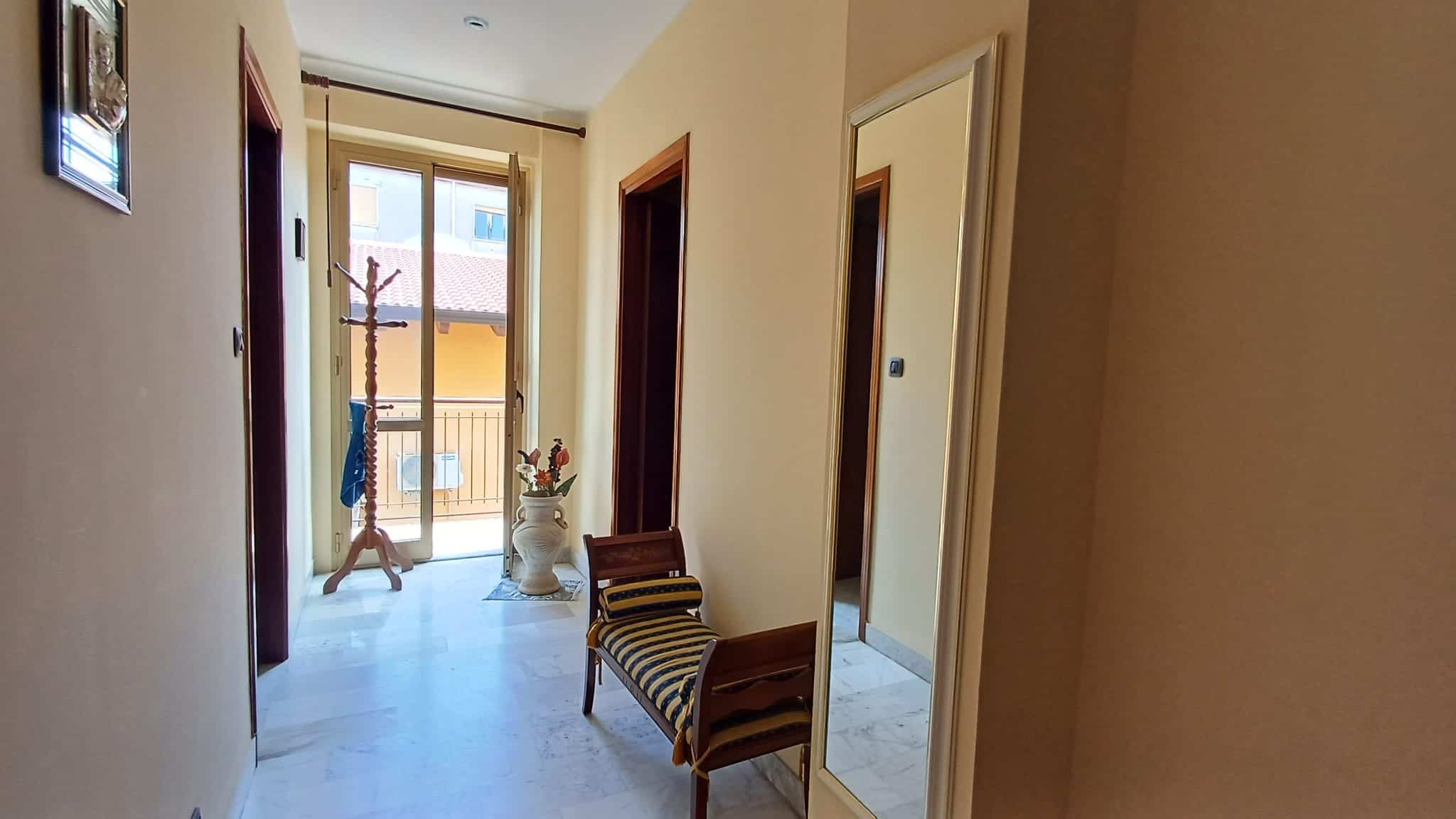 Elegante ingresso di casa con porta finestra che apre su un balcone, arredato con panca e decorazioni floreali, illuminato da luce naturale, ideale per ambienti residenziali di qualità.