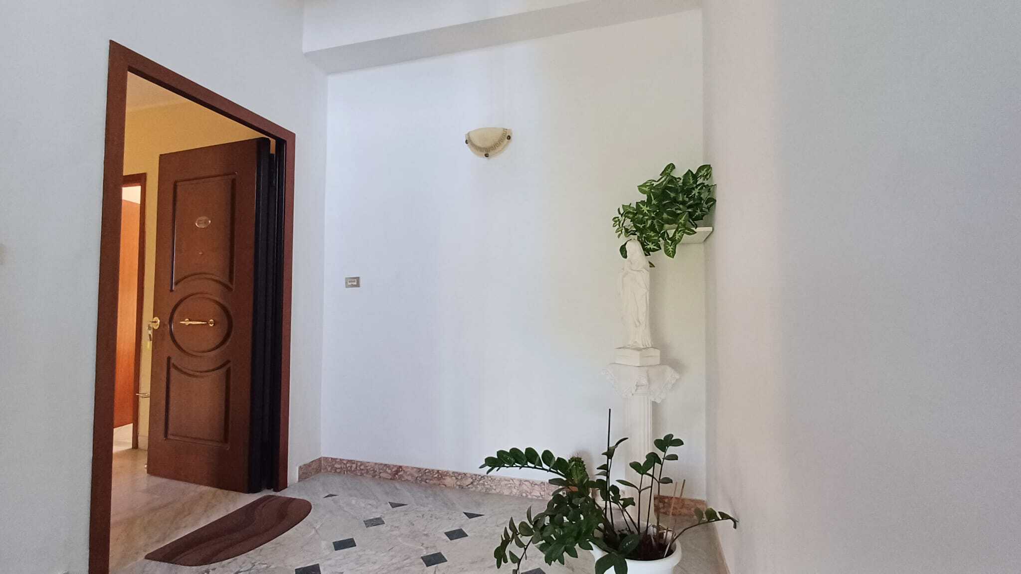 Porta d'ingresso in legno massiccio con dettagli decorativi, situata in un ingresso luminoso e spazioso con pareti bianche e piastrelle in marmo chiaro, ideale per appartamenti e case moderne.