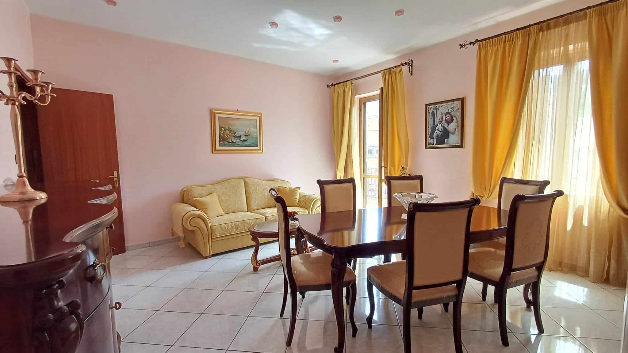 Elegante soggiorno con tavolo da pranzo, divano e grandi finestre con tende gialle, ideale per vivere momenti di relax e convivialità in un ambiente luminoso e accogliente.