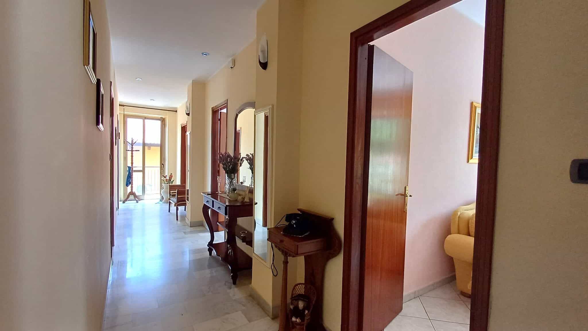 Ampia vista di un corridoio luminoso in un appartamento con pareti beige, arredi eleganti e accessori decorativi, che conduce a una zona giorno con balcone. Perfetto per chi cerca soluzioni immobiliar.