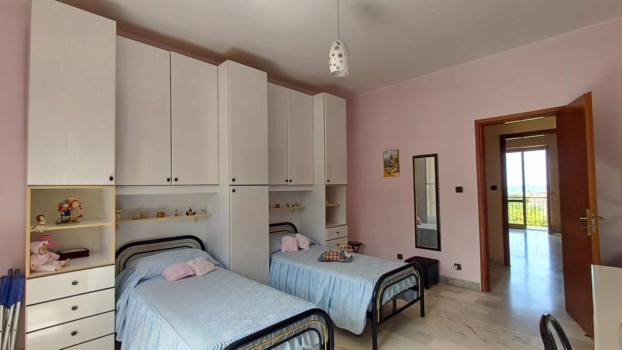 Letto doppio in camera con armadiature bianche e decorazioni colorate, vista sulla porta che conduce a un balcone con paesaggio naturale. Ideale per affitti o acquisti di immobili residenziali.