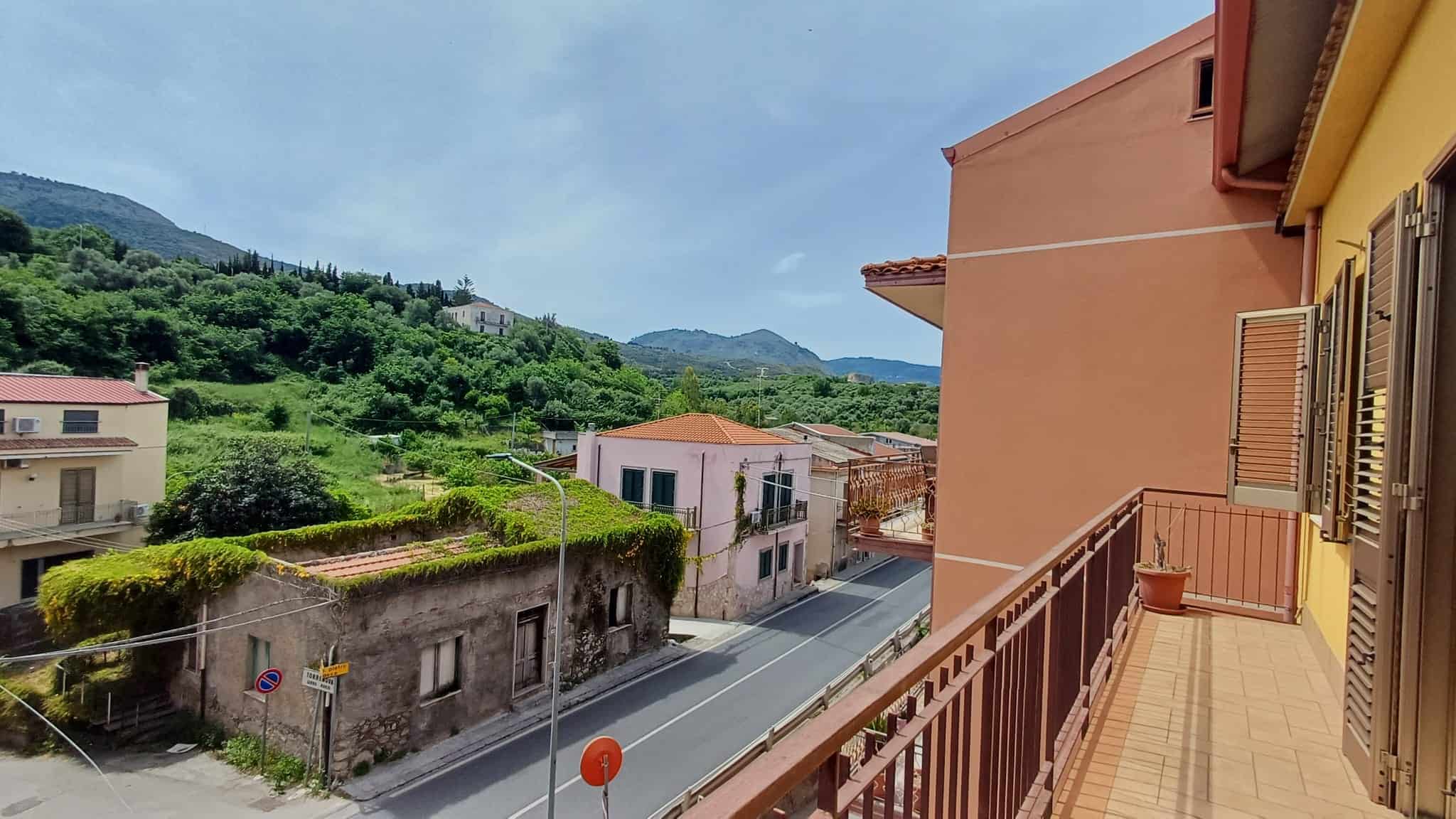 Balcone di un appartamento con vista su case e paesaggi collinari verdi, cielo sereno e montagne sullo sfondo, in un contesto residenziale tranquillo e pittoresco in Italia.