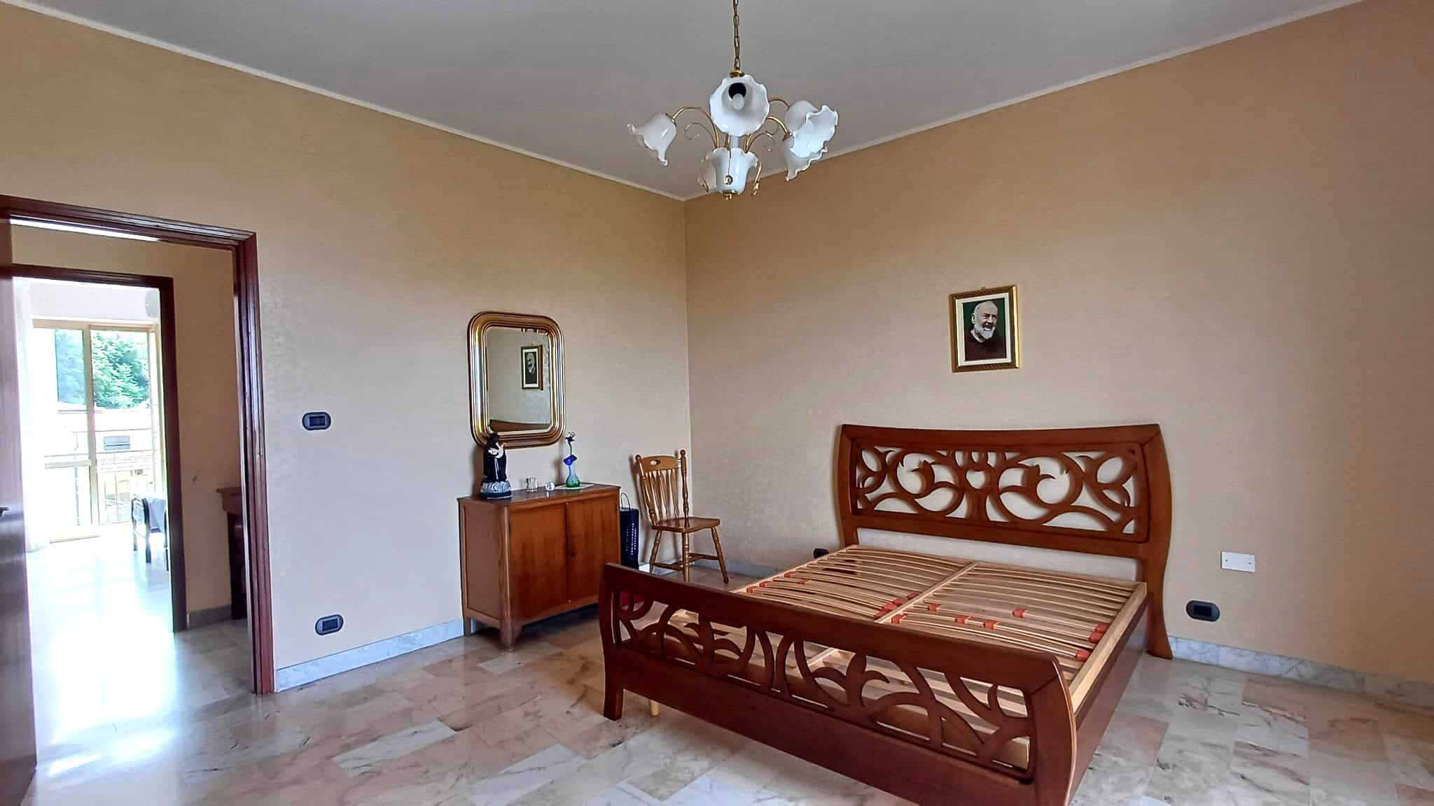 Letto matrimoniale in una camera spaziosa con pareti beige, arredi in legno e un quadro decorativo sopra la testata. La stanza è luminosa grazie alla luce naturale proveniente dalla porta aperta.