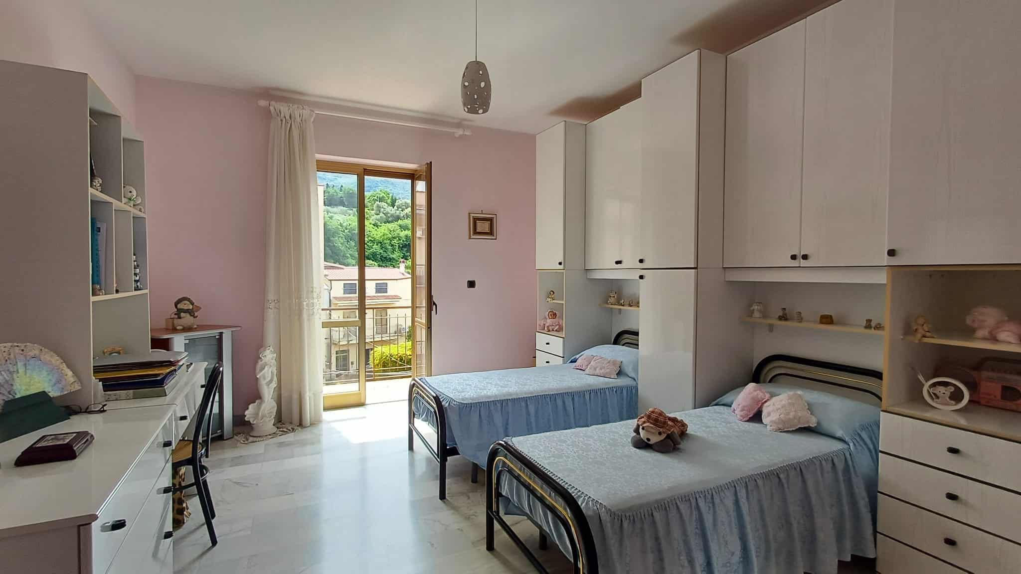 Letto singolo con biancheria in una camera luminosa e accogliente, con armadiature e accessori decorativi, vista su un balcone con paesaggio verde.