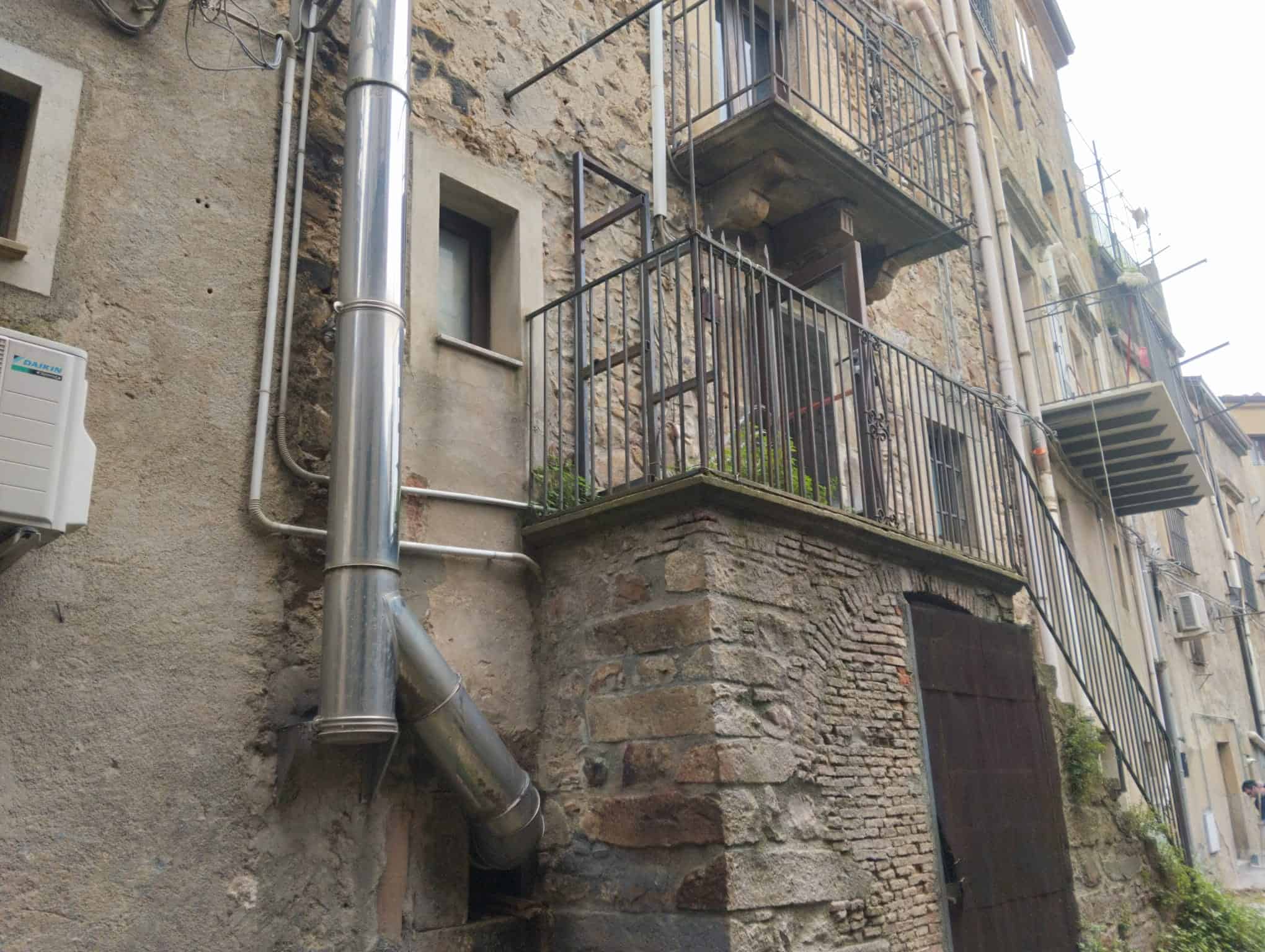 Vano esterno di un edificio storico con balcone in ferro battuto, pareti in pietra e tubature visibili, in un quartiere urbano di città italiane.
