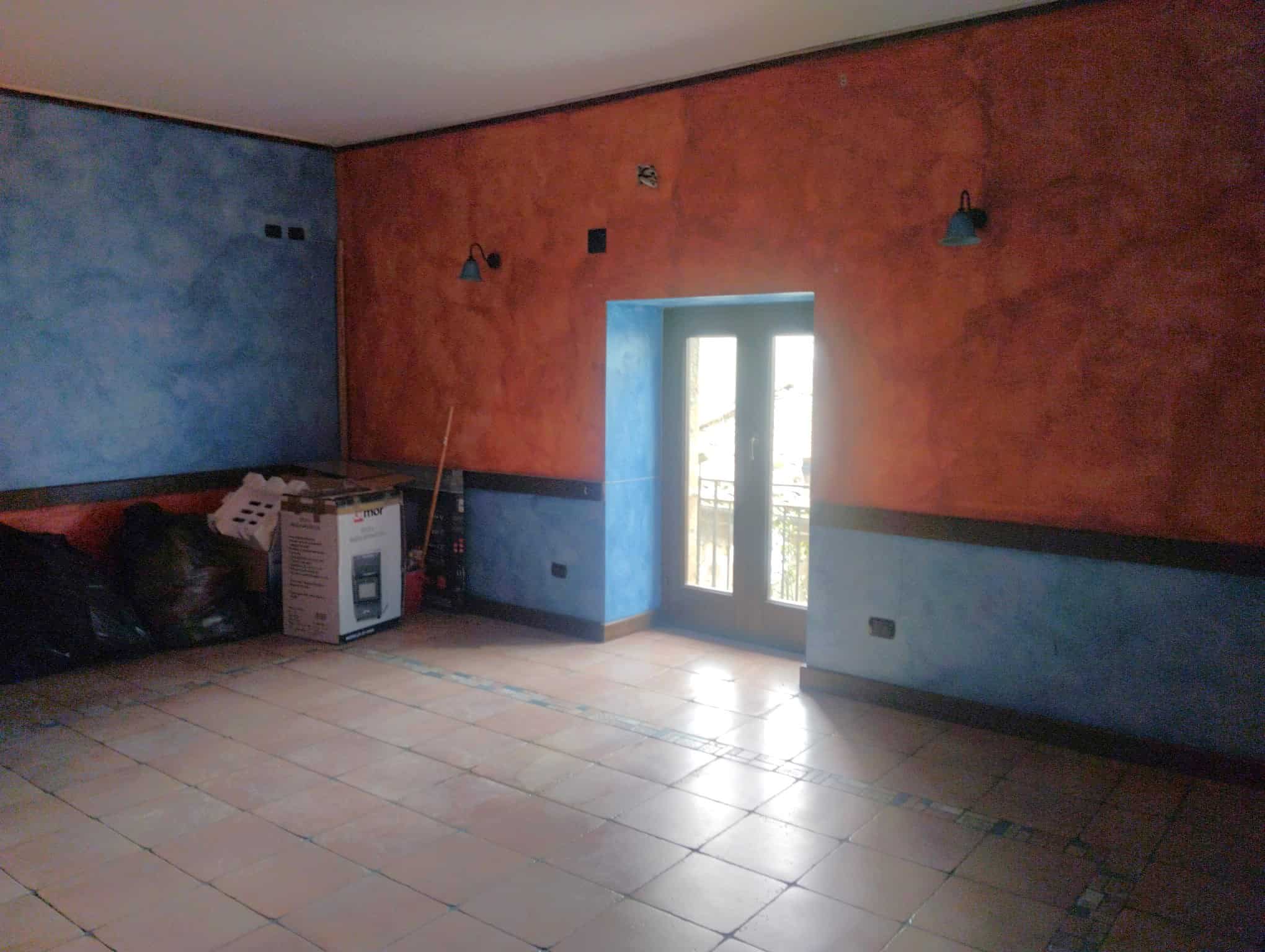 Aria luminosa e spaziosa in fase di ristrutturazione con pareti colorate in blu e rosso, porta finestra che permette l'accesso a un balcone, ideale per ambienti residenziali moderni e personalizzabili.