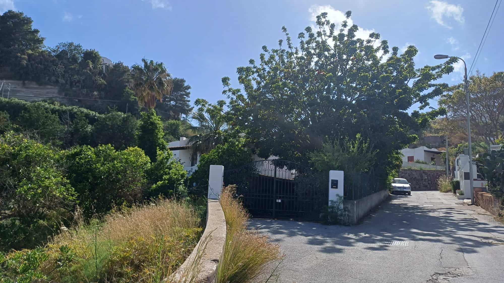 Ampia strada di campagna con ingresso in ferro battuto, circondata da vegetazione verde e alberi, sotto un cielo sereno e soleggiato, ideale per residenze tranquille e immersi nella natura.