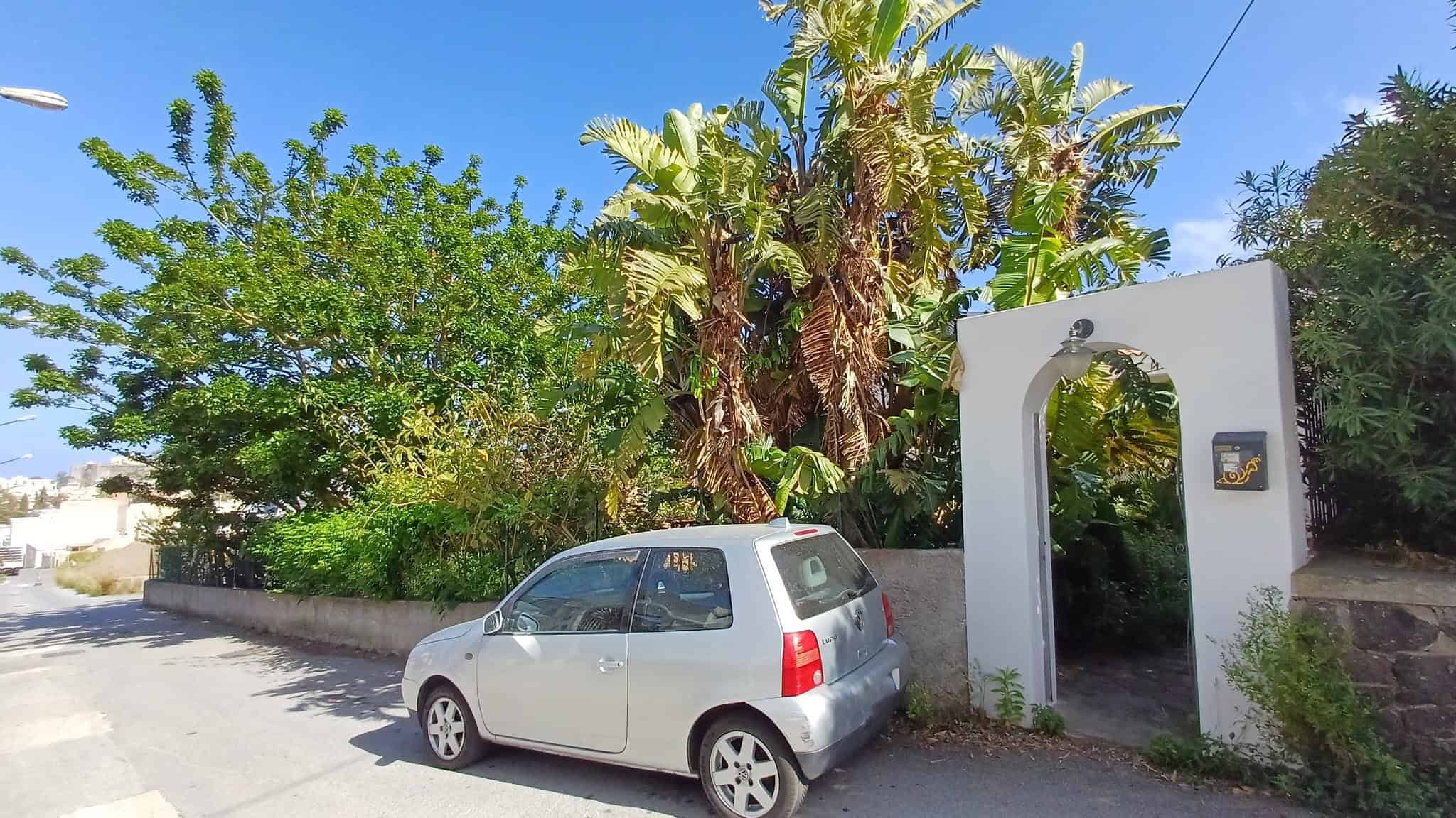 Auto compatta bianca parcheggiata su strada vicino a un ingresso con arco bianco, circondata da vegetazione lussureggiante e palme, in un ambiente residenziale soleggiato e tranquillo.