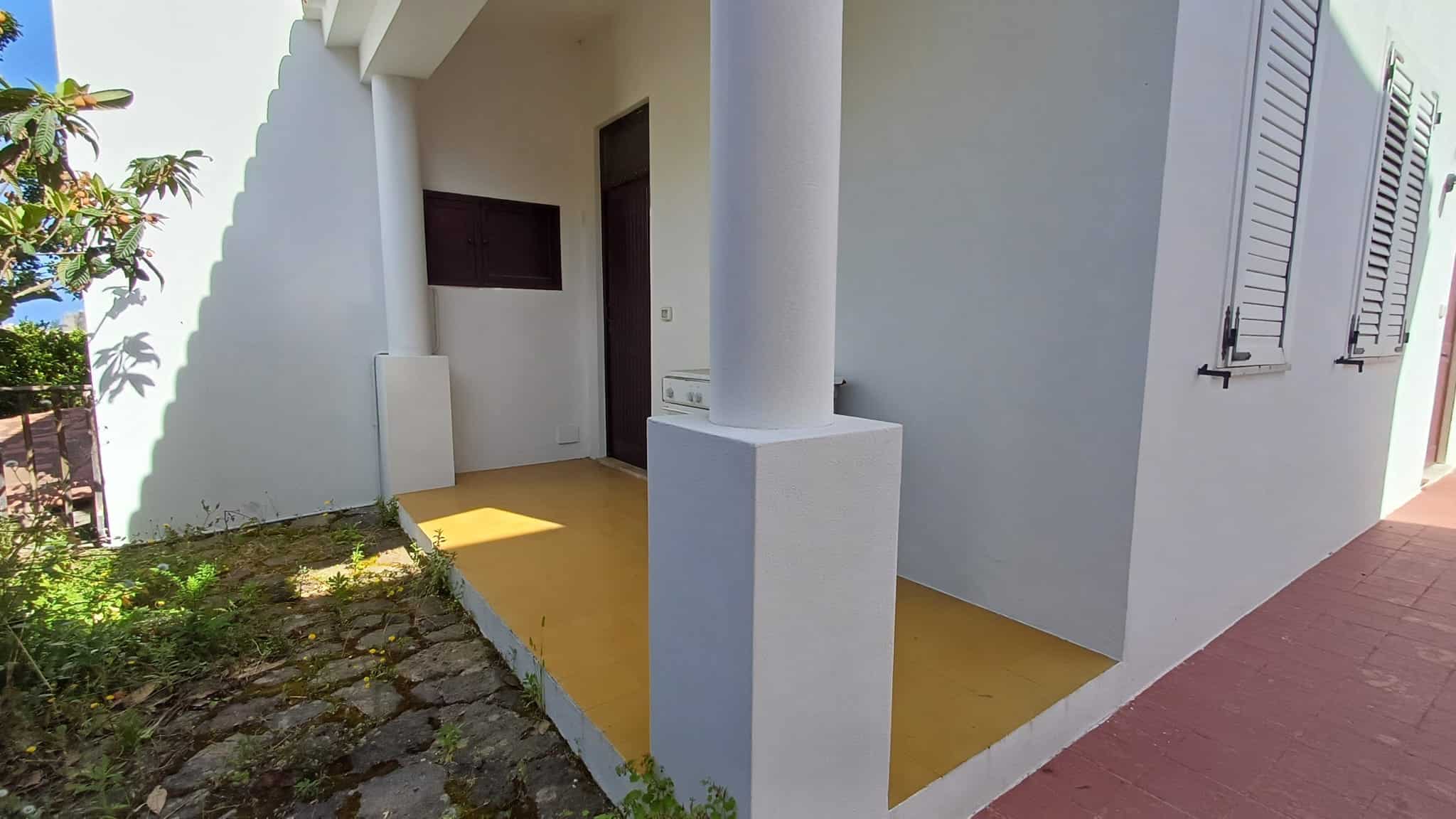 Ampia entrata di casa con portico bianco, colonne e pavimentazione gialla, situata in un contesto residenziale soleggiato con giardino e piante verdi, ideale per immobili in vendita o affitto.