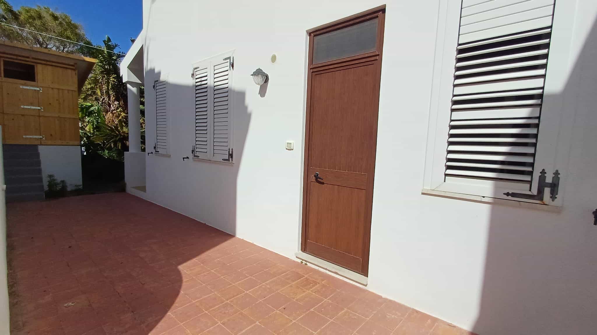 Ampia porta in legno e finestre con persiane in alluminio bianco su parete bianca, ingresso di una casa moderna con pavimentazione in piastrelle rosse.