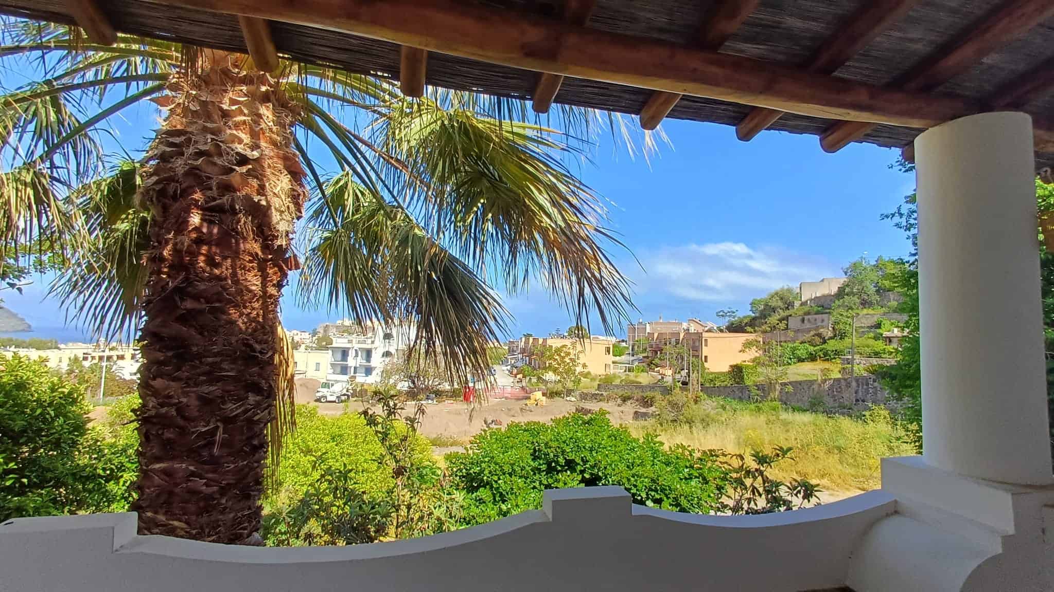 Aggiungi una palma e una vista panoramica di un paesaggio urbano e naturale dal balcone di una casa in stile mediterraneo, con cielo sereno e edifici sullo sfondo.