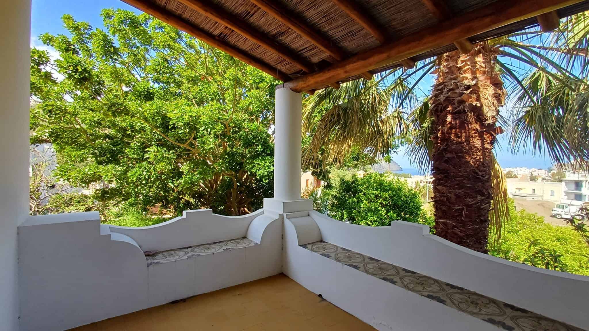 Ampio balcone con vista su alberi e palme, coperto da tetto in legno, ideale per relax e momenti all'aperto, in un contesto residenziale tranquillo e verdeggiante.