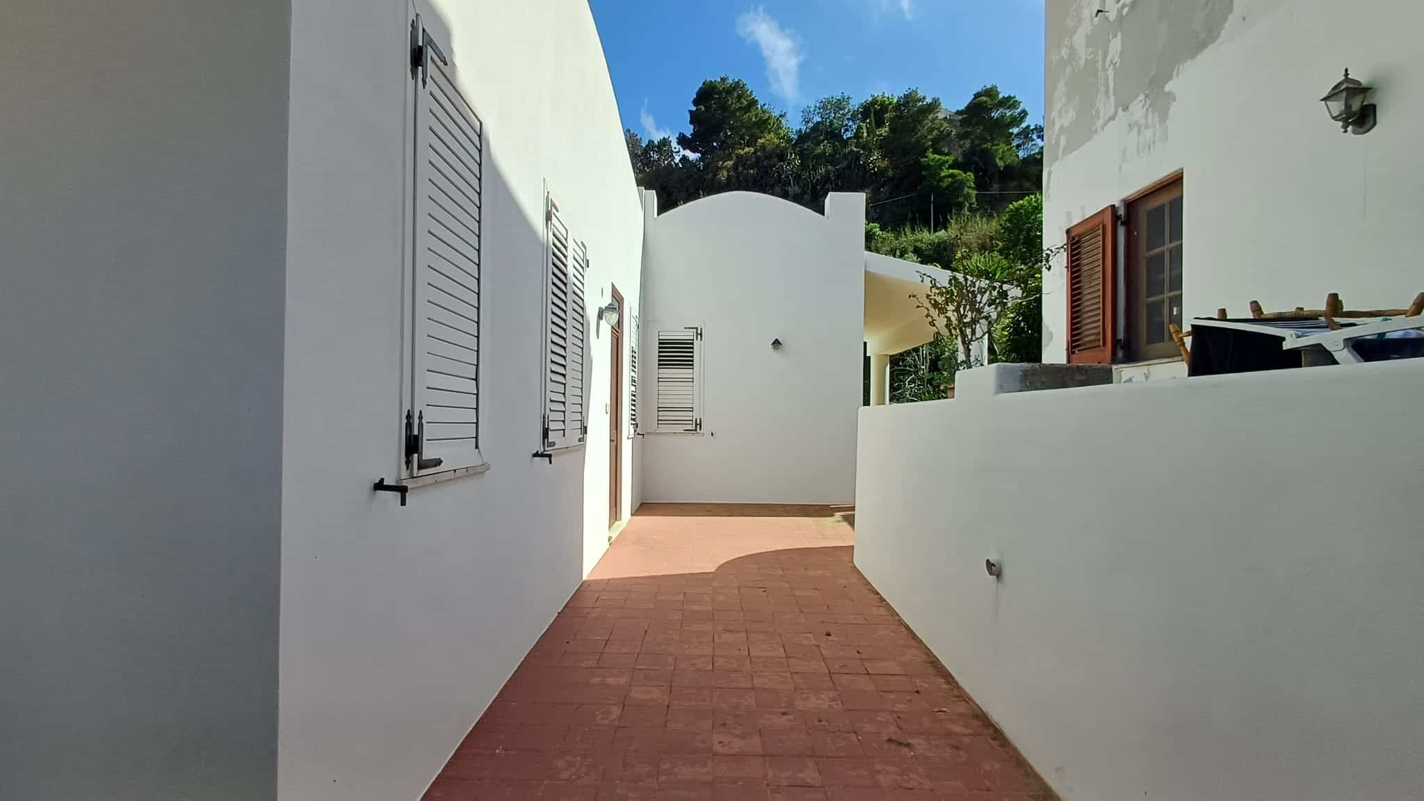 Elegante cortile esterno di una casa bianca con pavimentazione in terracotta, finestre con persiane e un ambiente tranquillo immerso nel verde, ideale per relax e convivialità.