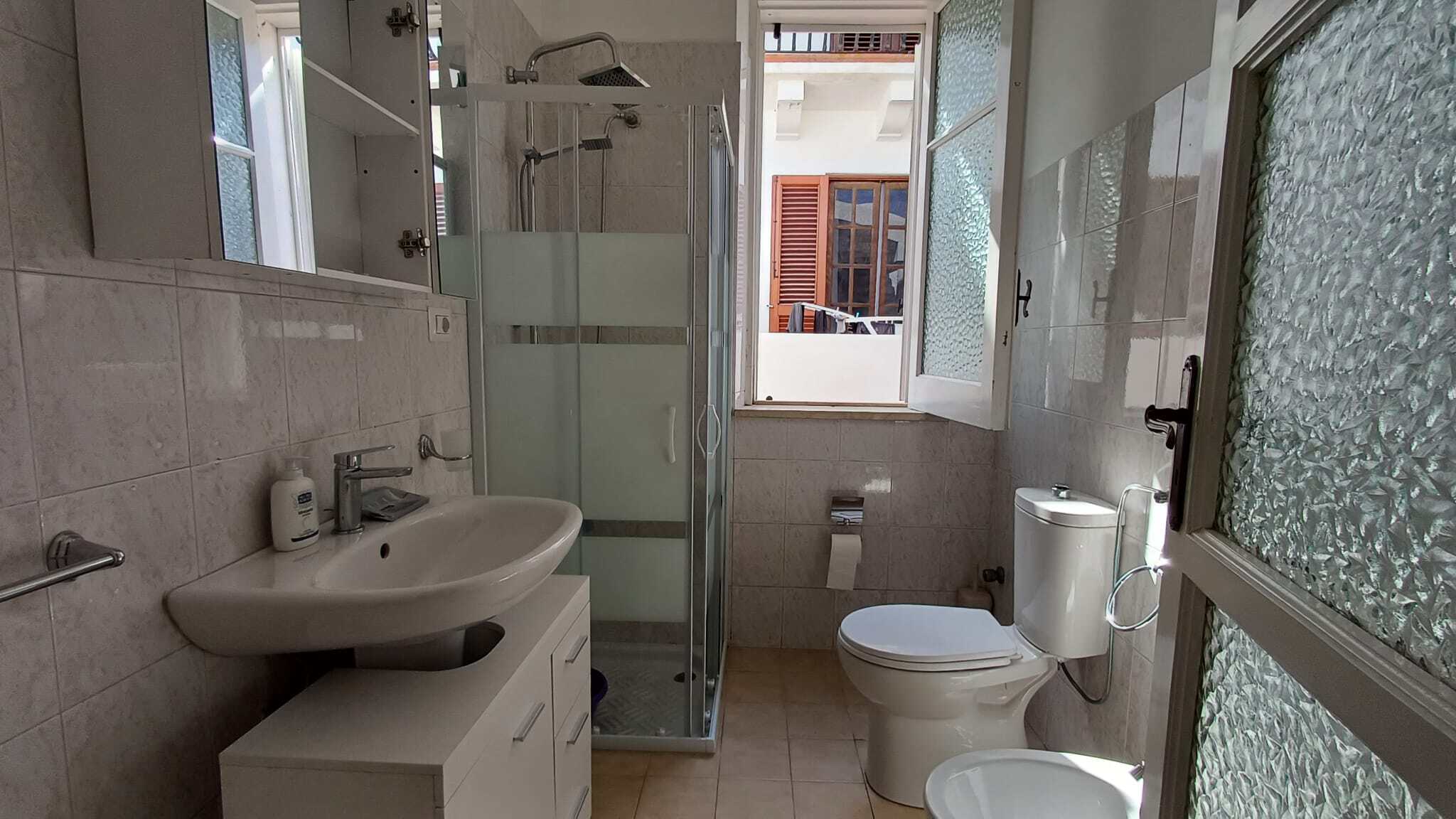Ampia stanza bagno moderna con doccia, WC e lavabo, illuminata da luce naturale attraverso finestra con persiane in legno, pareti e pavimenti in piastrelle chiare, ambiente pulito e funzionale.