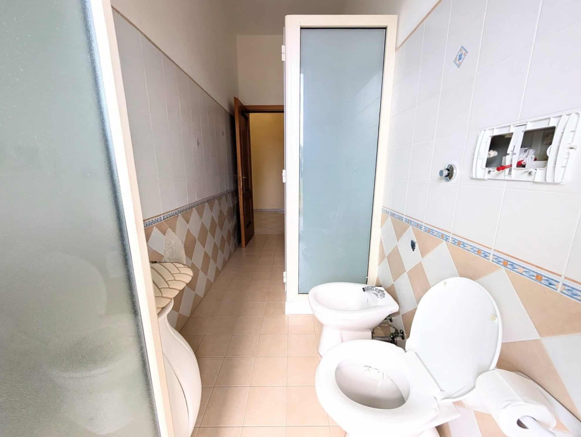 Bagno con sanitari e pareti rivestite di piastrelle beige e azzurre, porta in vetro e spazio funzionale, ideale per ambienti domestici o commerciali.