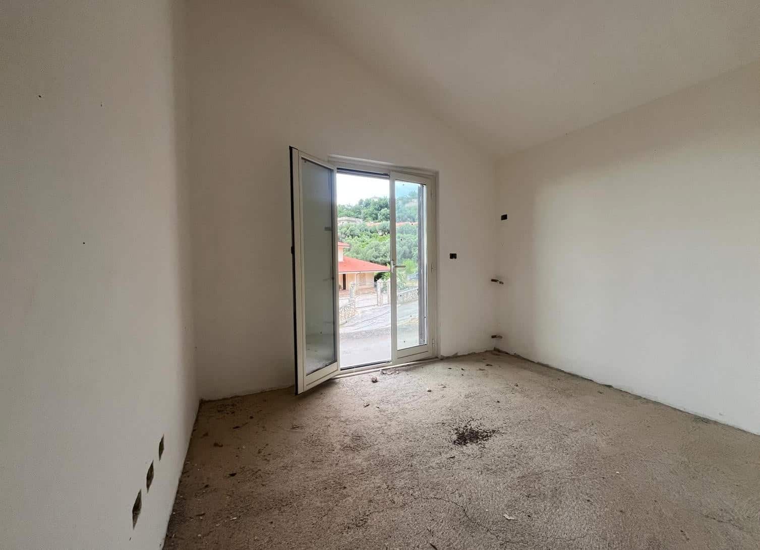 Stanza vuota in fase di ristrutturazione con porta finestra che dà su un balcone e vista su aree verdi, pronta per personalizzazione e arredo.