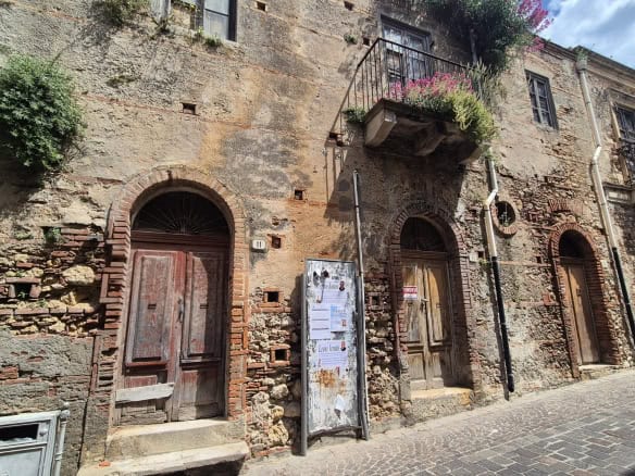 Antico edificio in pietra con porte in legno e balcone fiorito, situato in una strada acciottolata nel centro storico di un paese italiano. Perfetto esempio di architettura tradizionale e charme local.