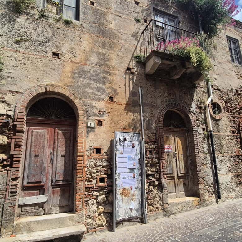 Antico edificio in pietra con porte in legno e balcone fiorito, situato in una strada acciottolata nel centro storico di un paese italiano. Perfetto esempio di architettura tradizionale e charme local.
