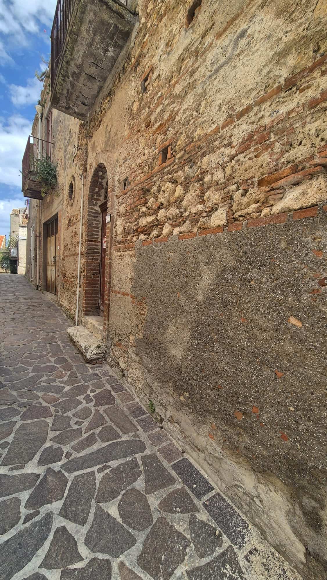 Antico edificio in pietra con pareti in mattoni e strada in pietra, in un quartiere storico italiano, con un cielo azzurro e alcune piante sul balcone.