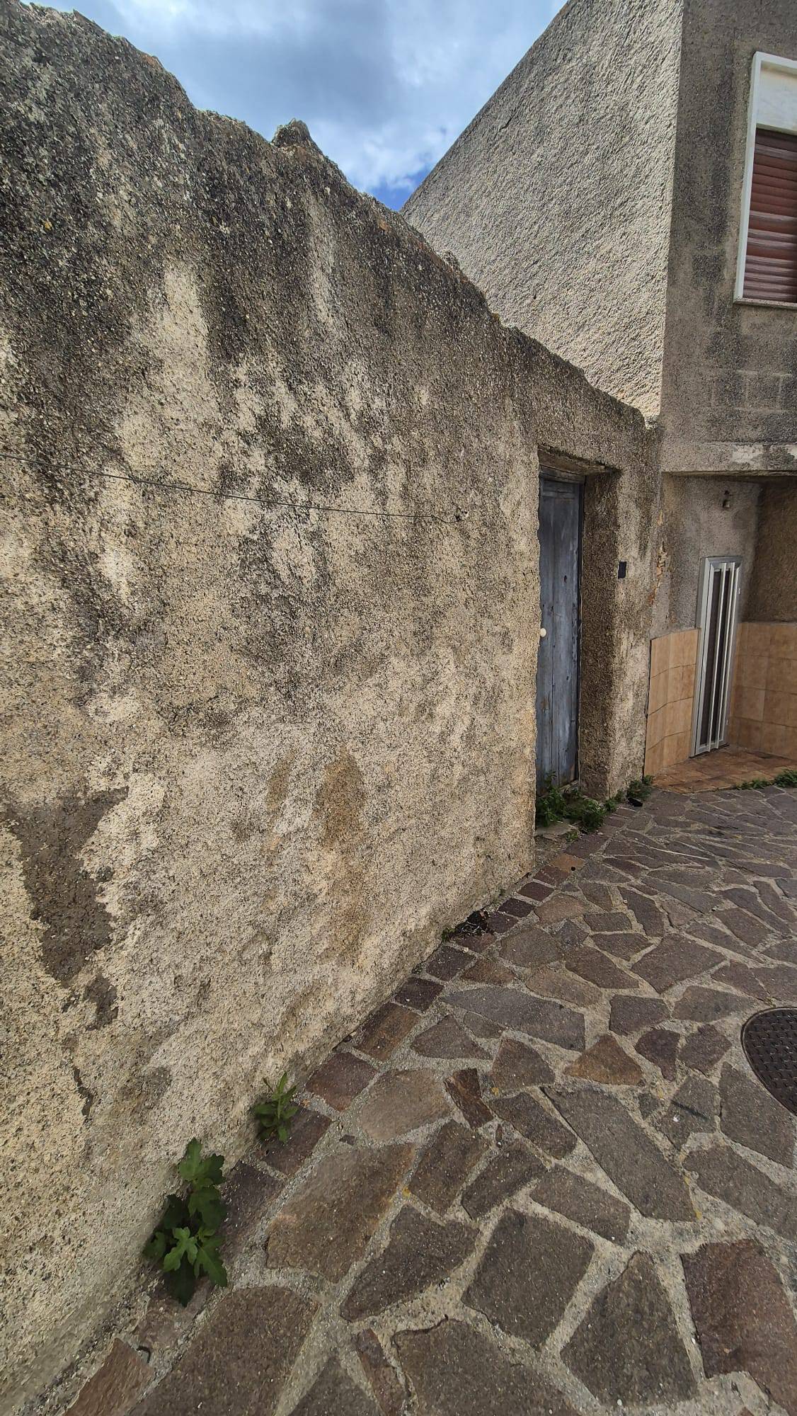 1. Muro esterno in pietra e cemento con porta e finestra, pavimentazione in pietra, in un contesto urbano storico.