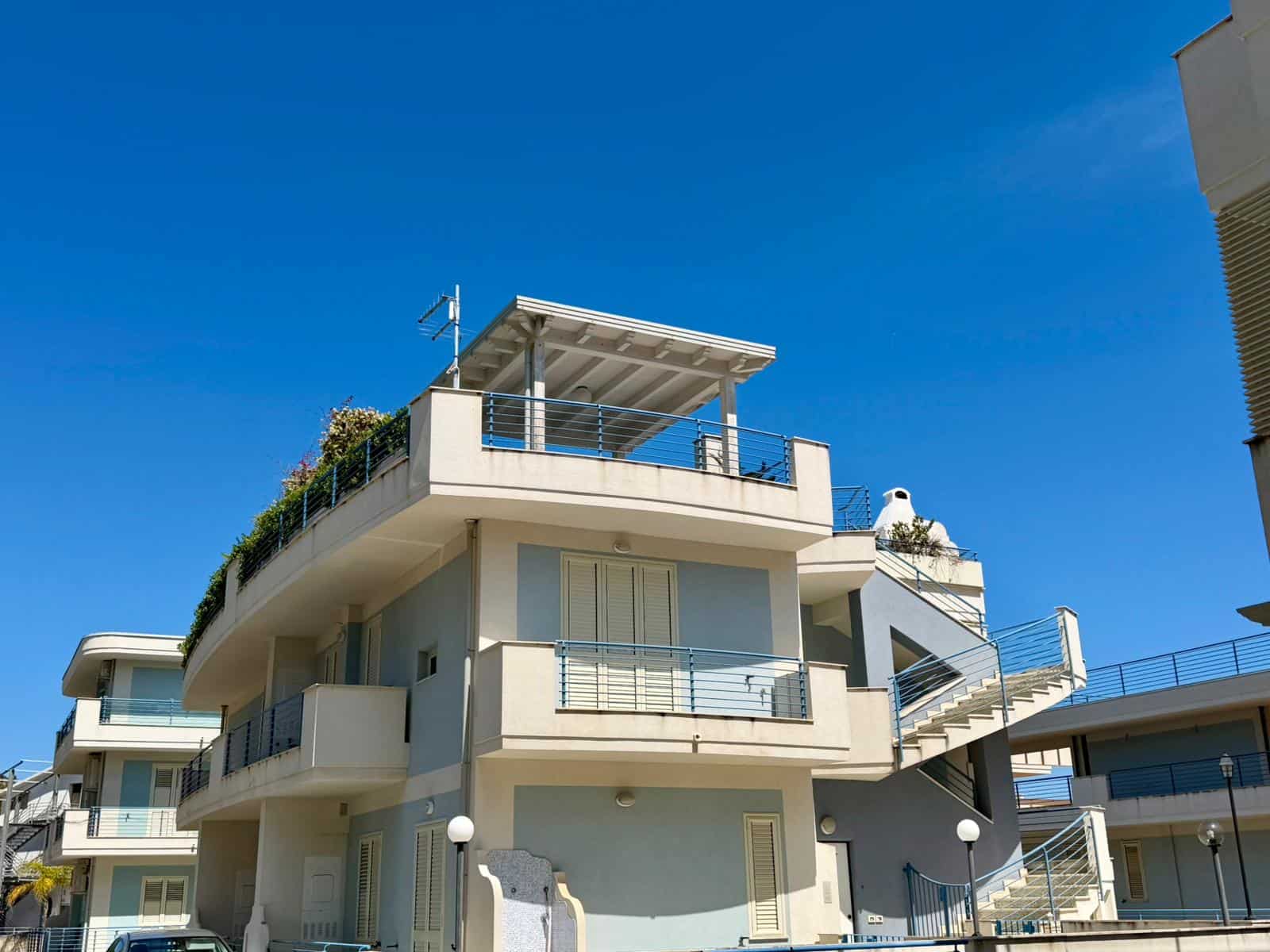 Balcone panoramico con vista sul mare, edificio moderno in stile contemporaneo, facciata bianca e balconi con ringhiere in metallo, cielo azzurro, zona residenziale di lusso, NextCasa.