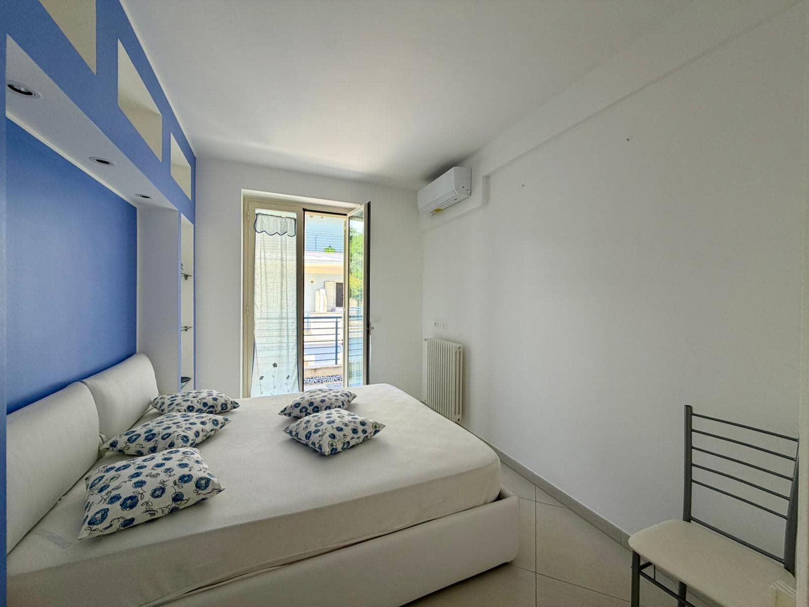 Ampia camera da letto moderna con pareti bianche e dettagli blu, letto matrimoniale con cuscini decorativi, aria condizionata e balcone con vista all'aperto, ideale per soggiorni confortevoli.