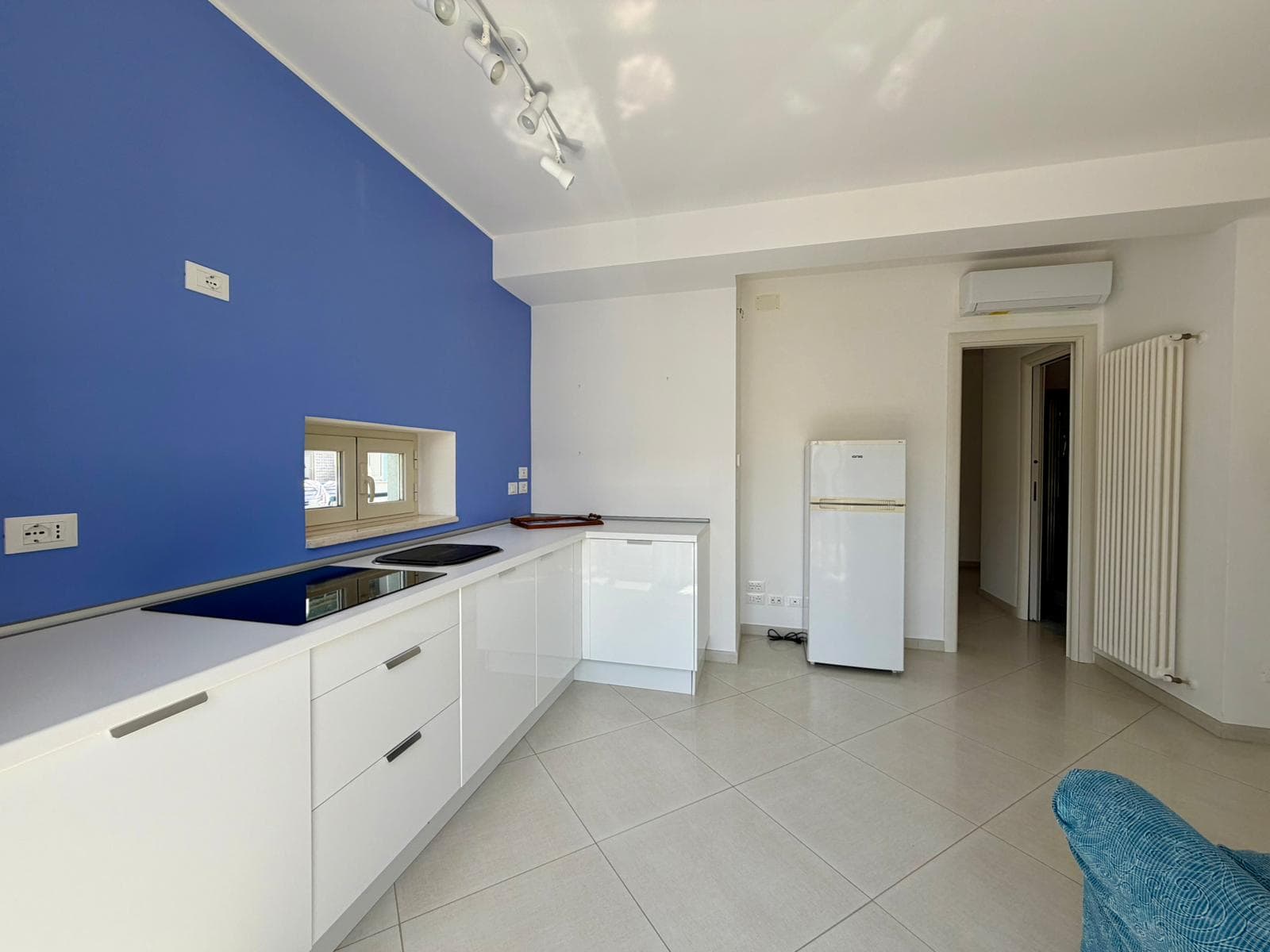 Lavagna di cucina moderna con parete blu, mobili bianchi, piano cottura a induzione, frigorifero e radiatore, ambiente luminoso e spazioso, ideale per case di design e ristrutturazioni.