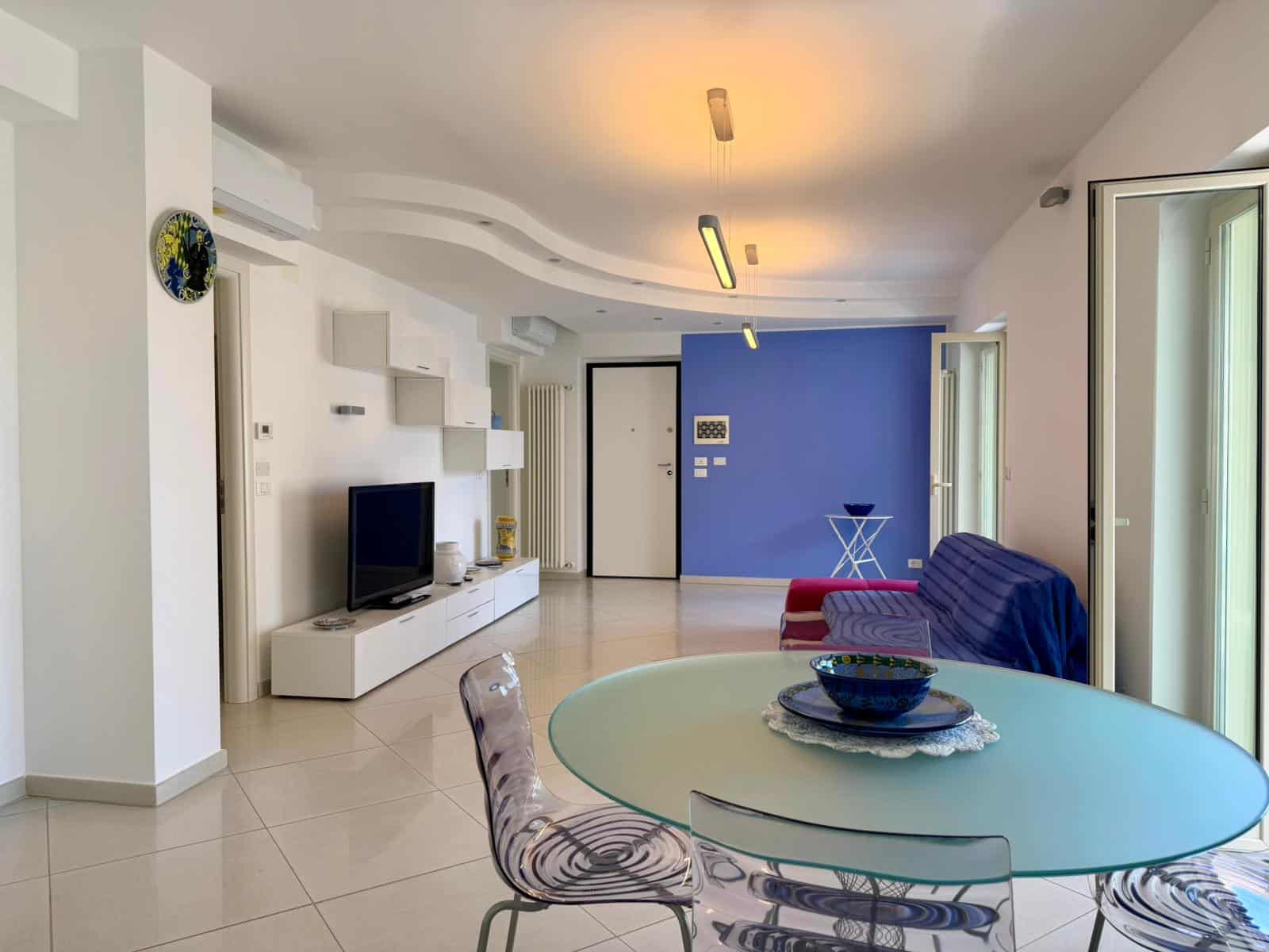 Ampia zona living moderna con pareti bianche, parete blu accentuata, divano rosso, tavolo rotondo in vetro e sedie eleganti, grandi finestre che lasciano entrare molta luce naturale, ideale per appart.