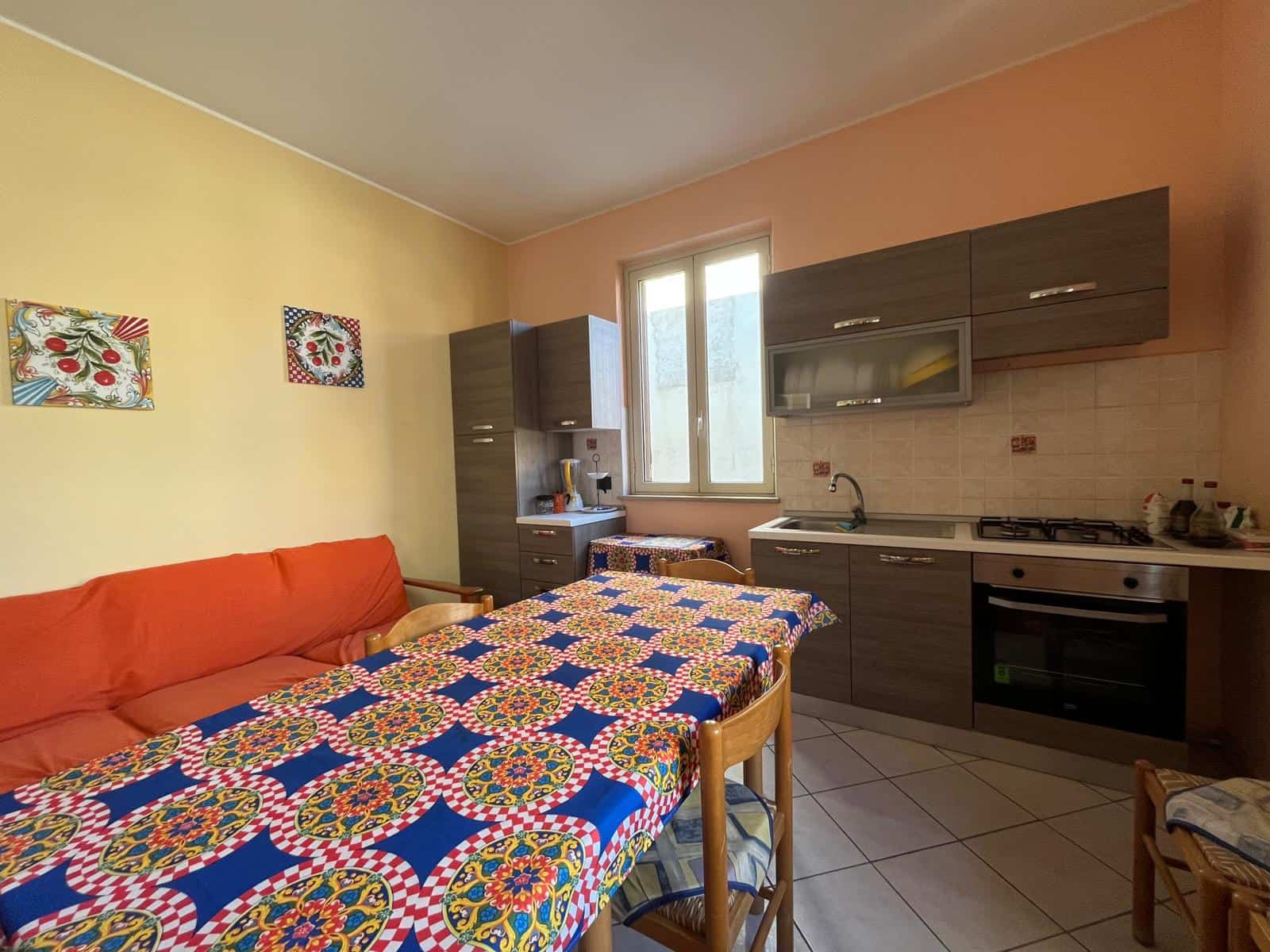 Un angolo cucina e zona pranzo in un appartamento con pareti colorate, tavolo con tovaglia decorata, cucina moderna con elettrodomestici e finestra che lascia entrare luce naturale.