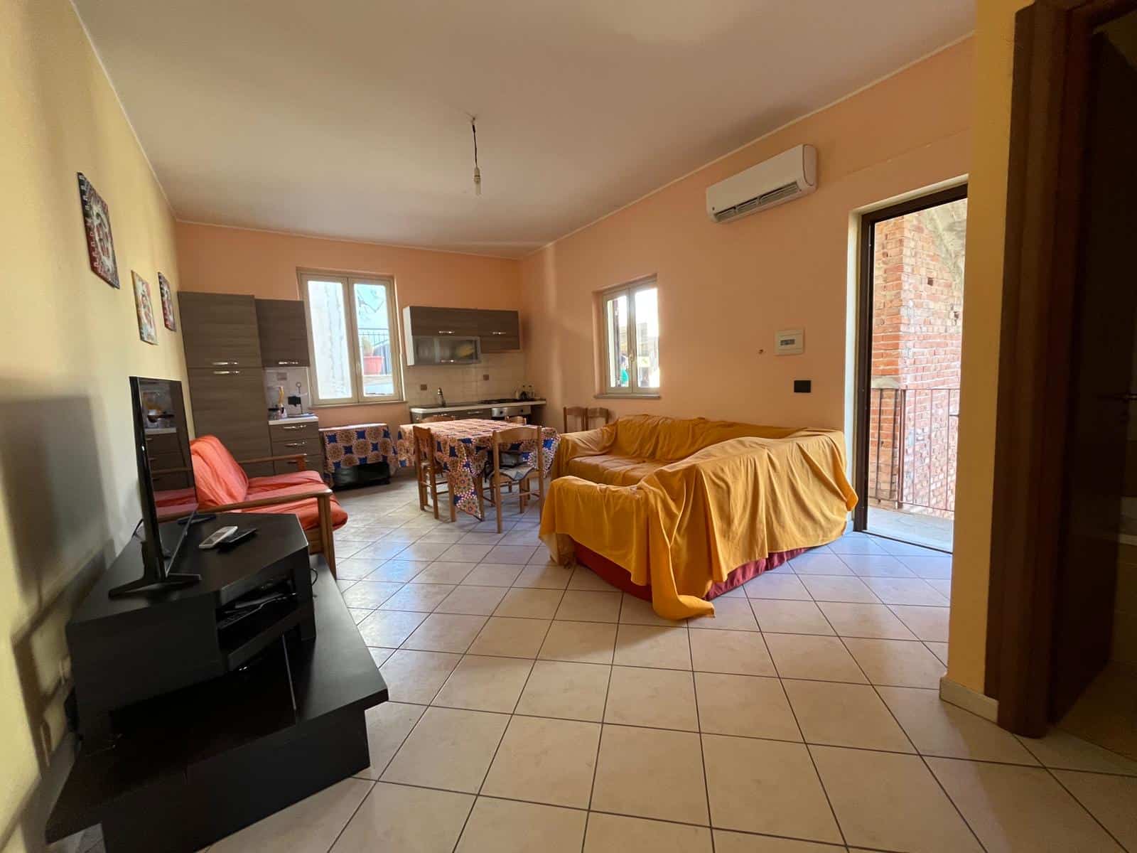 Interno di un soggiorno luminoso con divano, cucina compatta e zona pranzo, arredamento moderno e accessori funzionali, ideale per vivere in comfort e stile.