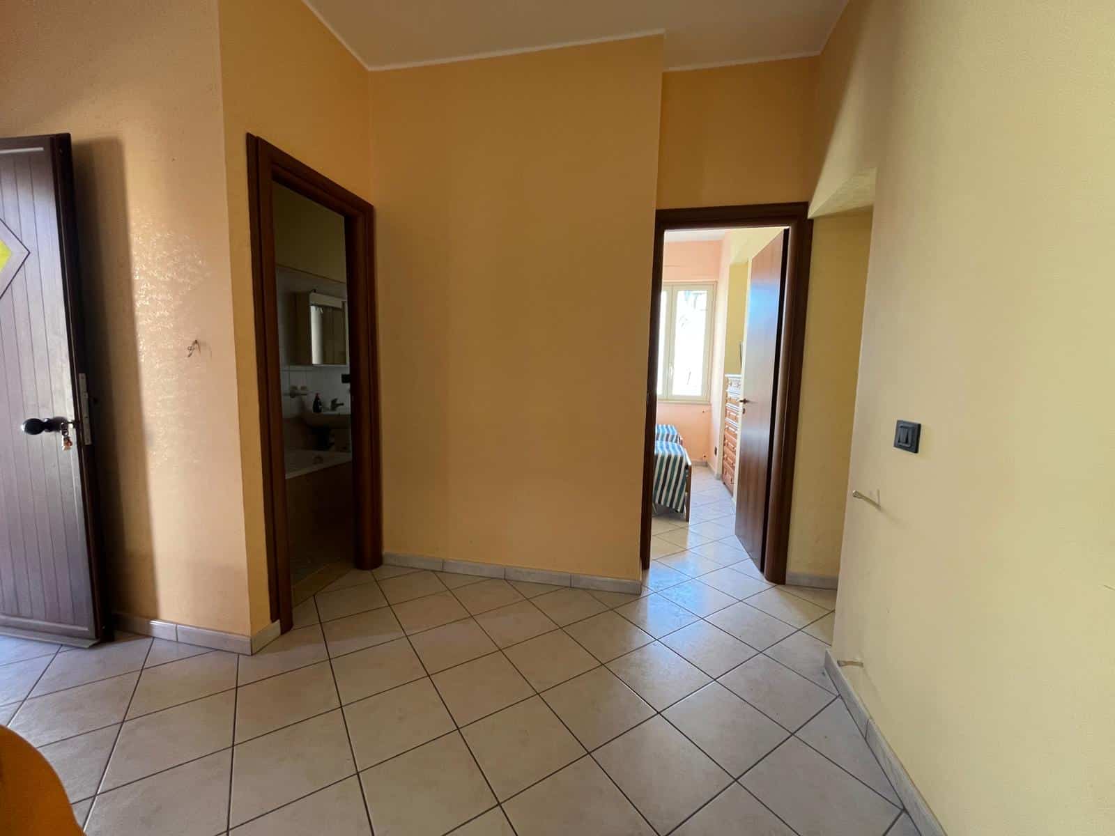 Interno di un appartamento con pareti beige, porte in legno e pavimento in piastrelle, vista verso le stanze e la luce naturale proveniente dalle finestre.