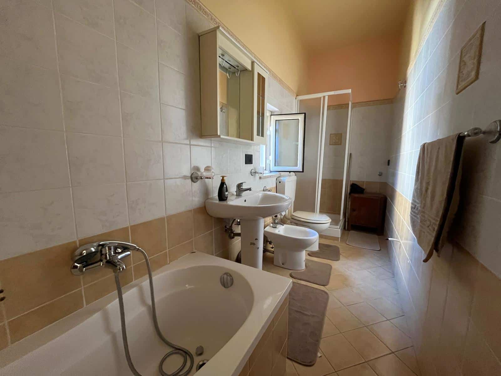 Bagno spazioso con vasca, lavabo e WC, illuminato da luce naturale attraverso una finestra aperta, con arredi semplici e funzionali, ideale per comfort e praticità.